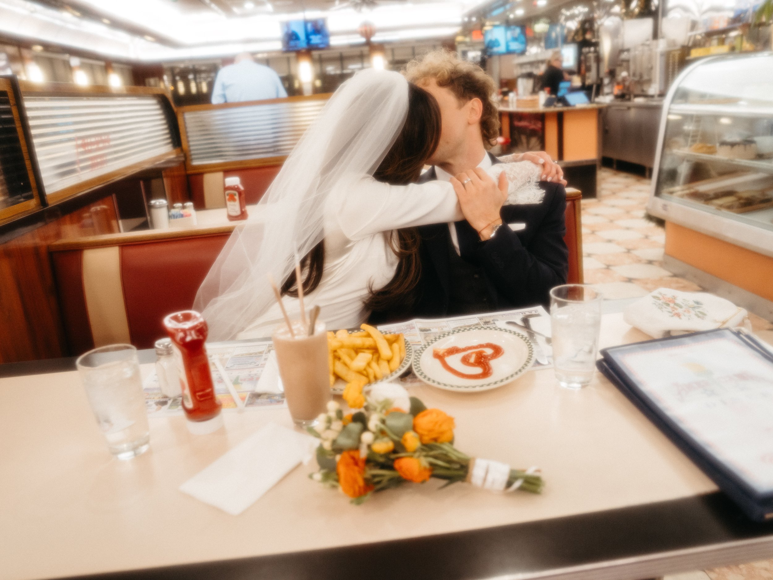 A bride and groom sitting at a table in a diner, sharing a kiss. The bride is wearing a white wedding dress and veil, and the groom is dressed in a dark suit. The table has fries, a milkshake, ketchup, drinks, and a flower bouquet.