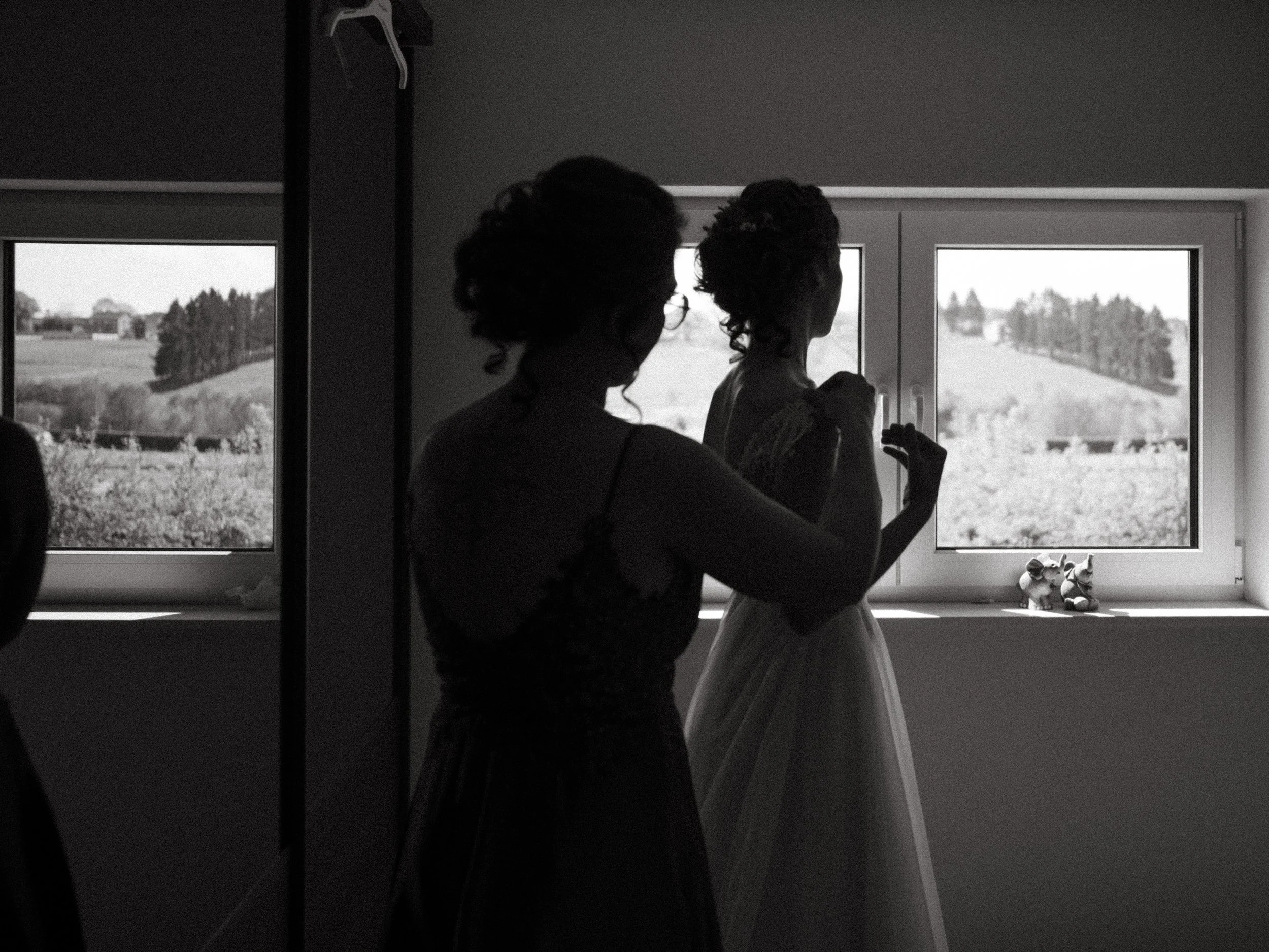 Black and white photo of two women, one in a dress, standing by a window with landscape view, possibly preparing for a wedding.