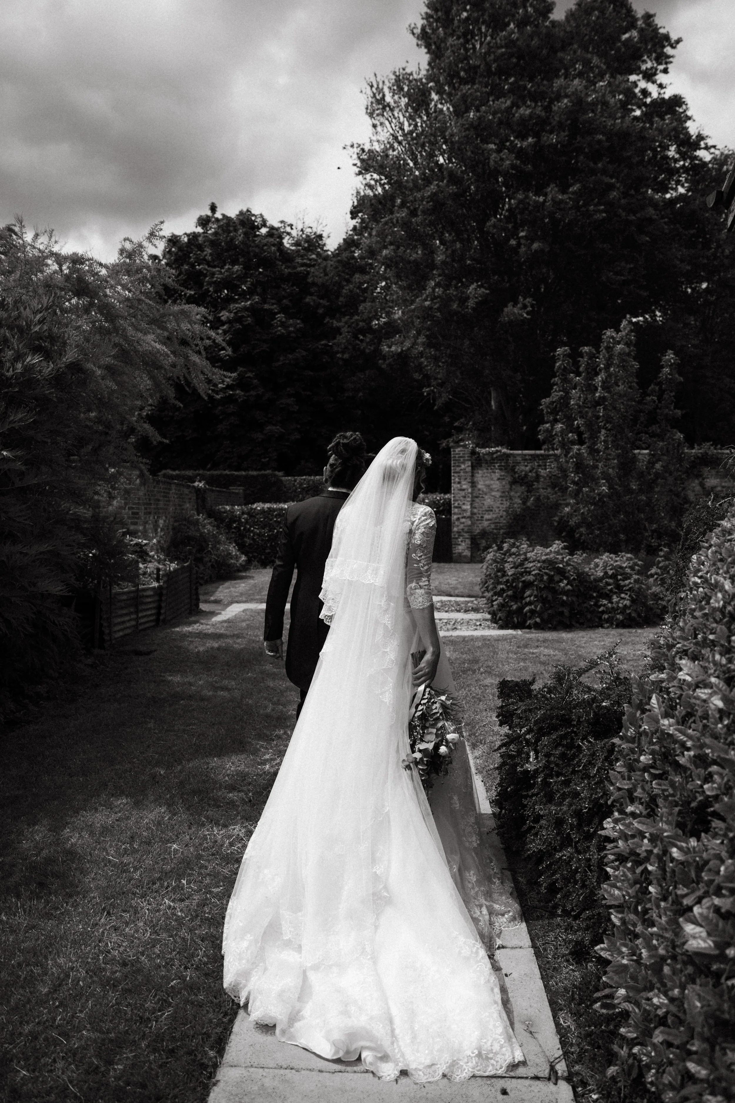 A black and white photo of a bride and groom walking together outdoors, with the bride holding a bouquet and wearing a long dress with a veil, and the groom in a suit.