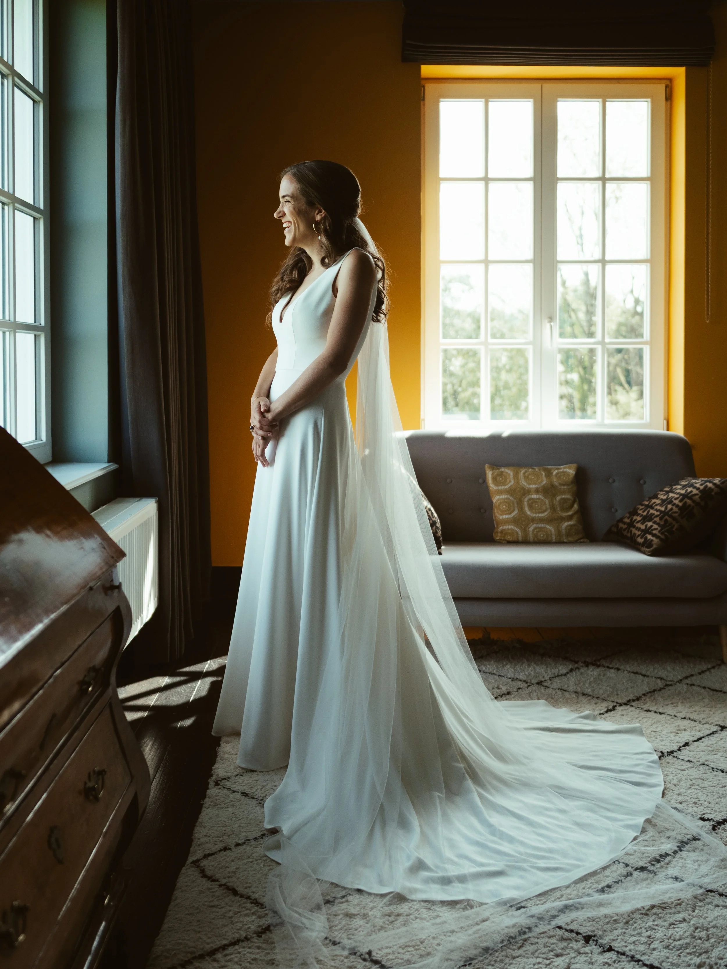 A bride in a white wedding dress standing by a window, smiling, with sunlight illuminating her face and dress inside a cozy room.