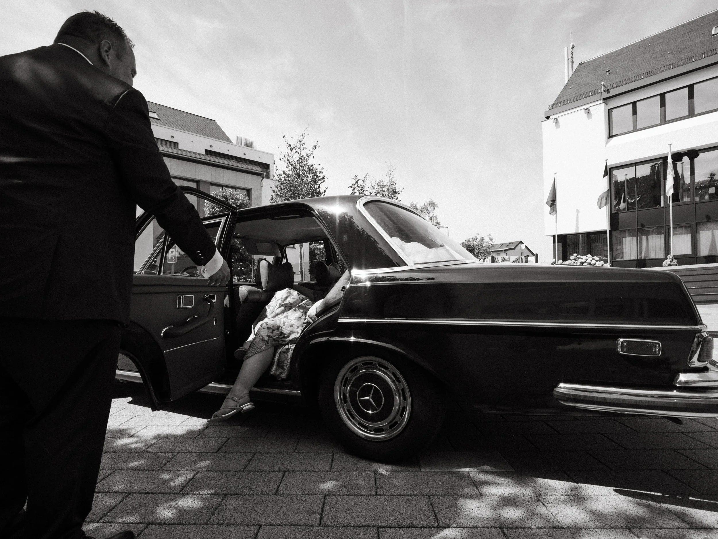 A man in a suit opening the door of a black vintage car as a woman lies inside, with her legs outside the car. The scene takes place outside a modern building with flags and trees in the background, captured in black and white.