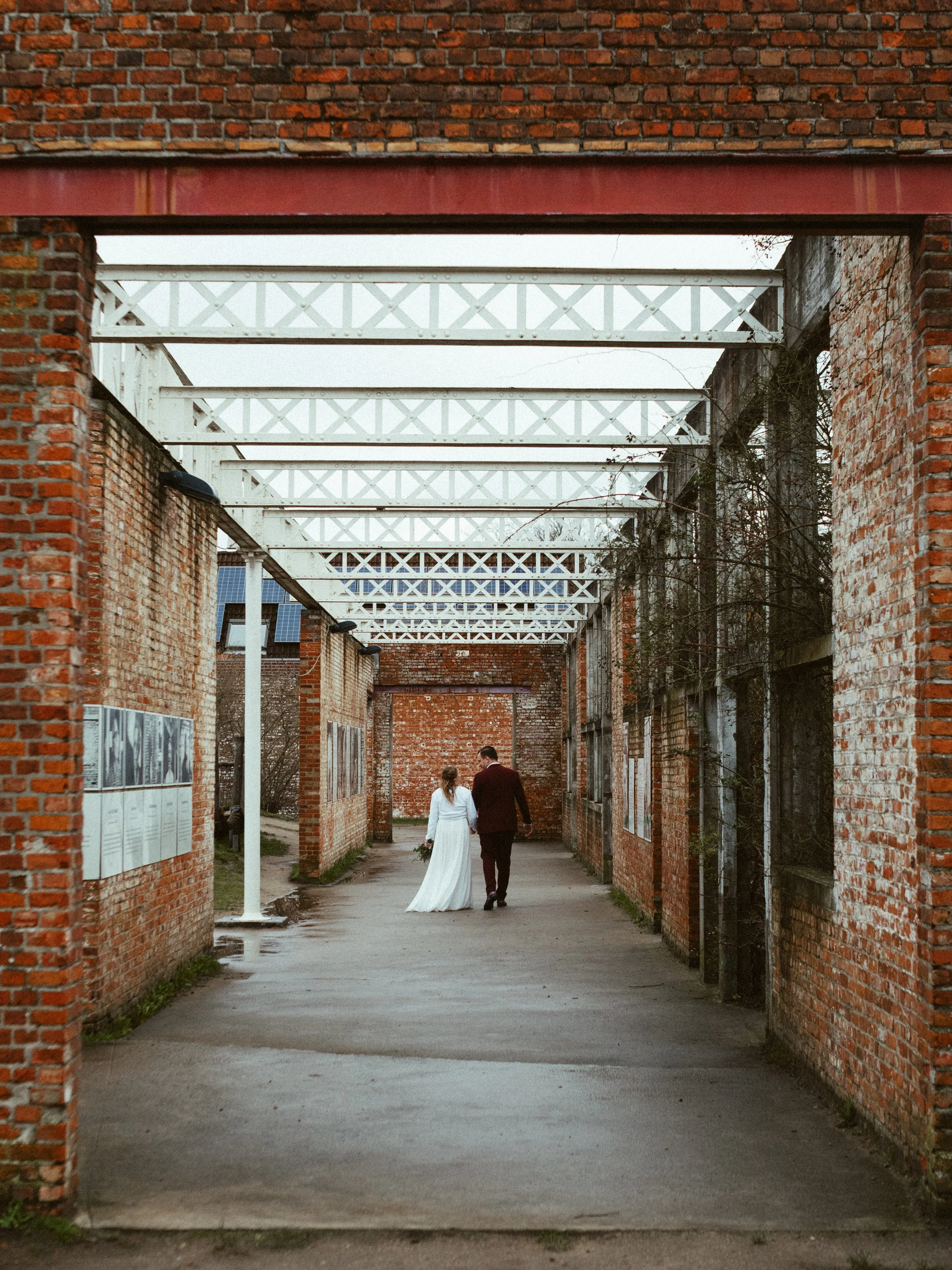 A bride and groom walking hand in hand through a brick alleyway with a metal roof structure overhead.