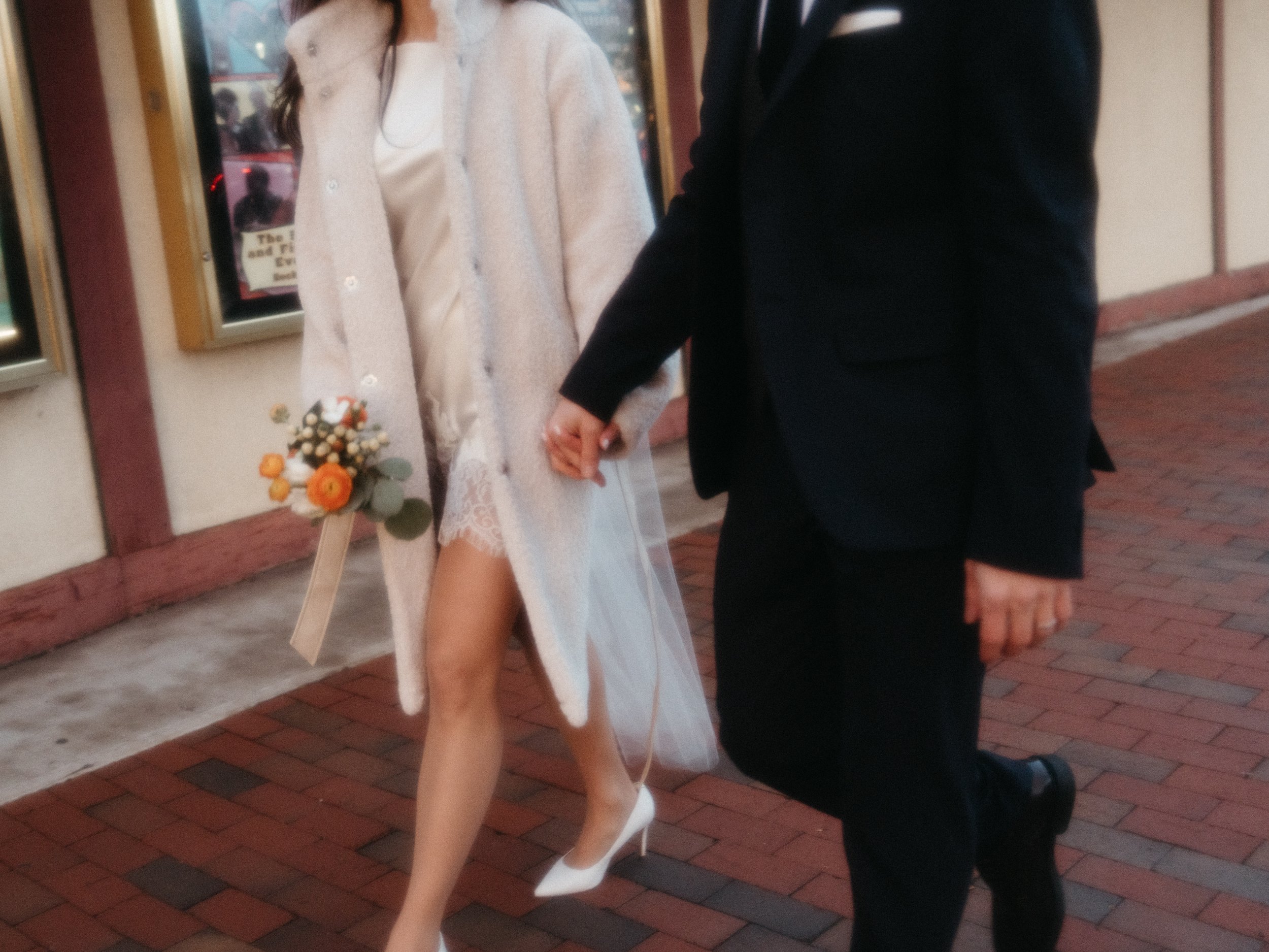 A person dressed in white high heels, a white dress, and a beige coat holding a bouquet of flowers is walking hand in hand with a person in a black suit on a brick sidewalk.