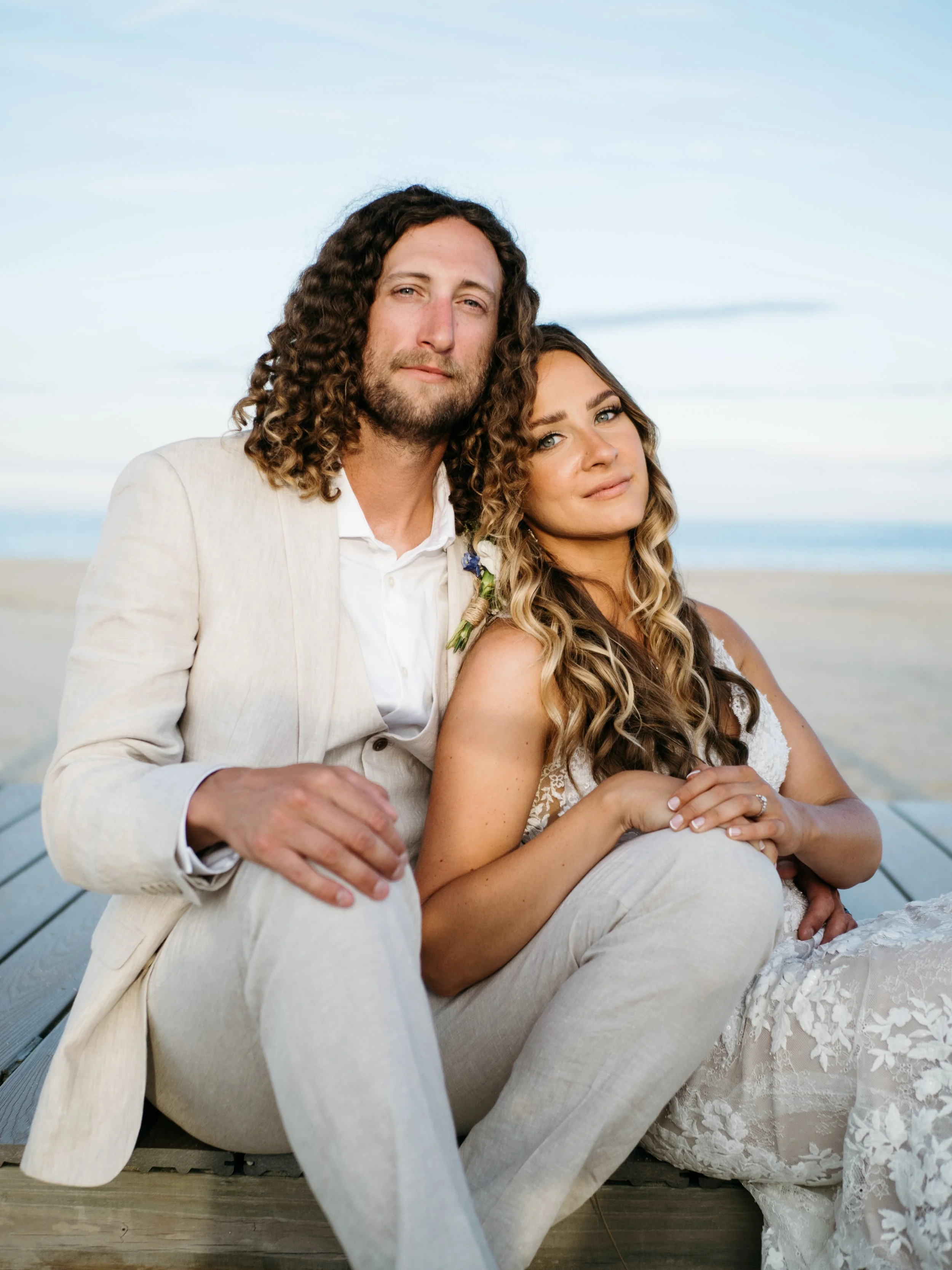 A couple sitting on a wooden dock at the beach during sunset. The man has long curly hair and a beard, wearing a cream-colored suit, and the woman has long wavy hair, wearing a white lace dress. They are sitting close together, with the woman leaning into the man.