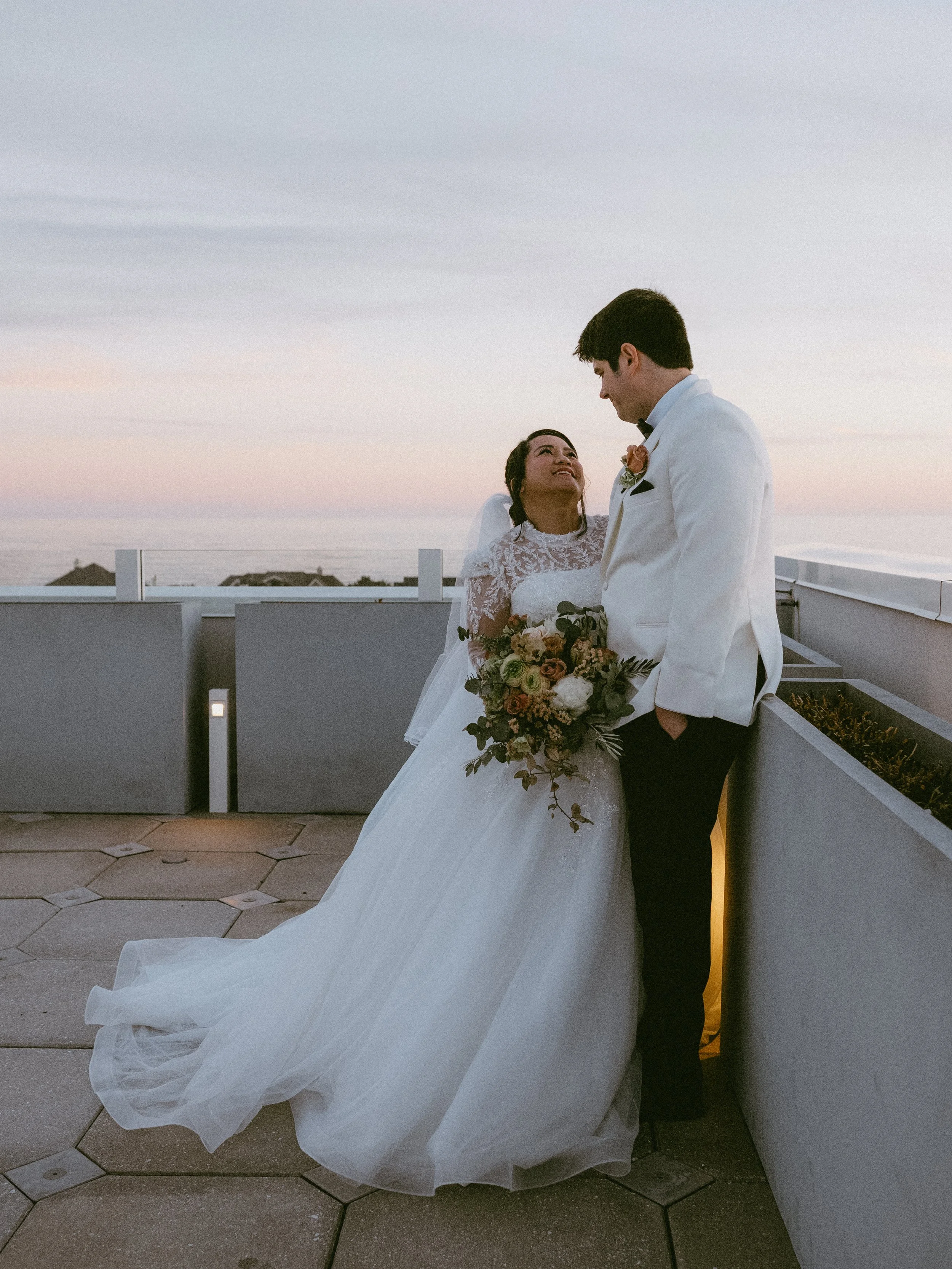 Bride and groom standing close together on rooftop at sunset, with the ocean in the background. The bride is holding a bouquet and looking up at the groom, who is dressed in a white tuxedo jacket with black pants.