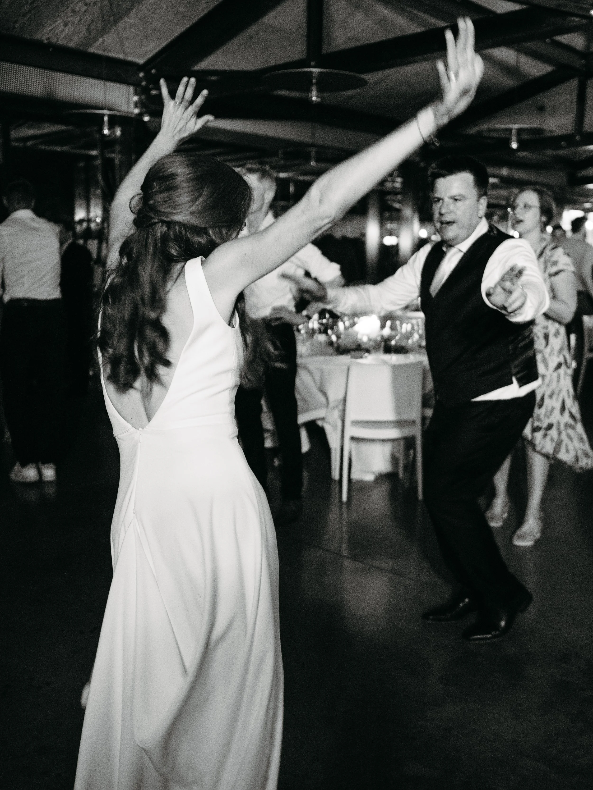 A woman in a white dress and a man in a tuxedo dance together at a celebration event, with other guests in the background.