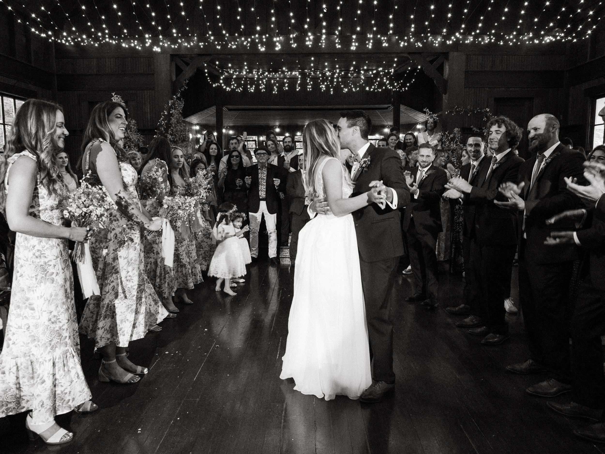 Black-and-white photo of a wedding dance with bride and groom at the center, surrounded by guests in formal attire, under string lights in a rustic indoor venue.