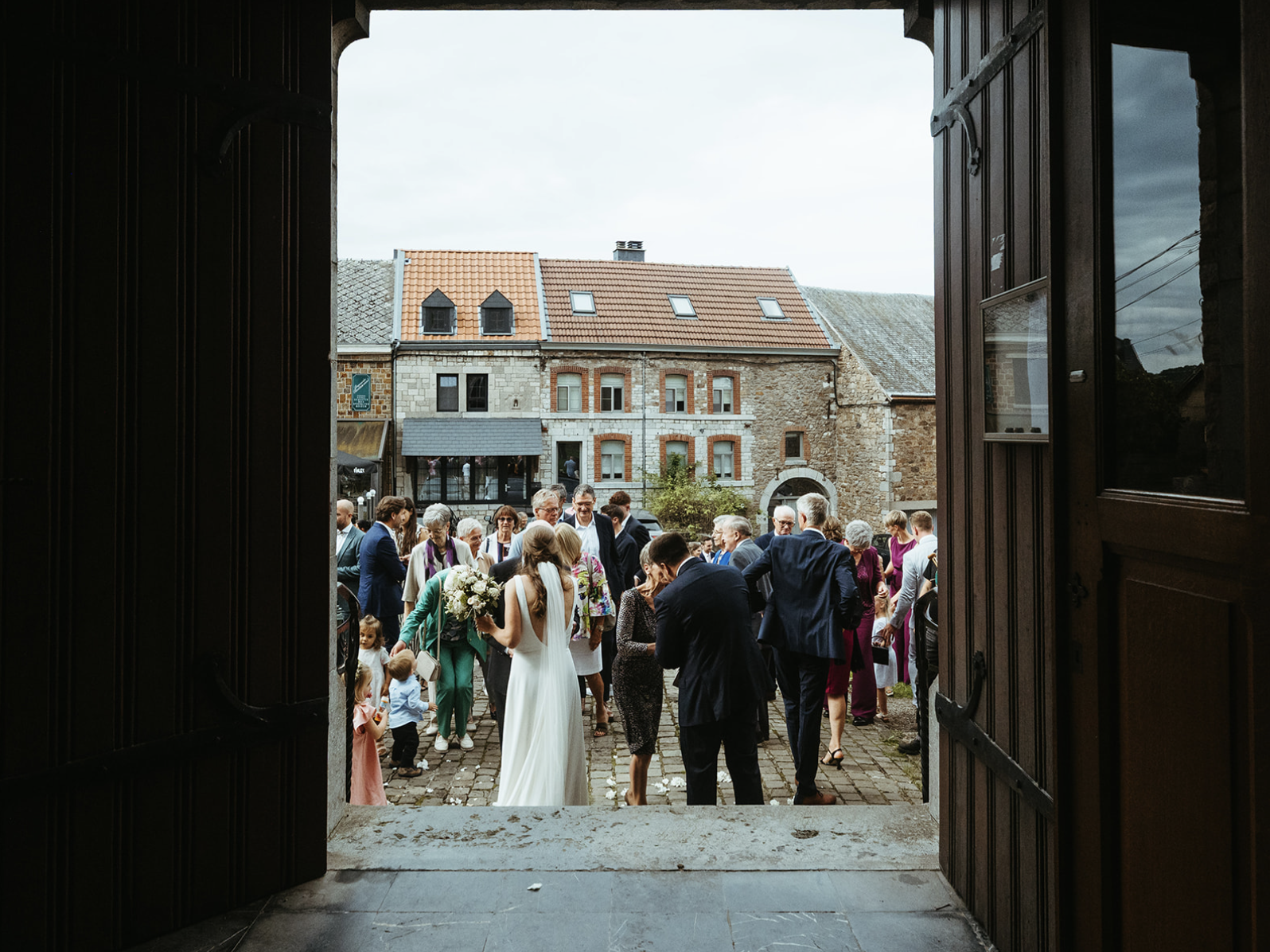 A gathering of people outside a building during a wedding celebration, viewed through an open doorway with dark wooden doors.