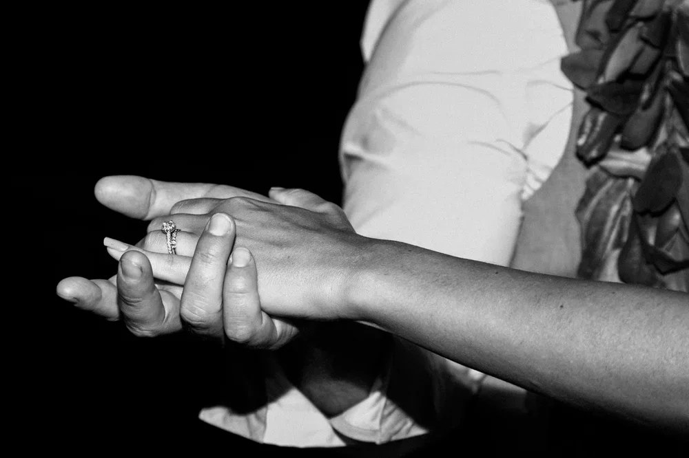 Close-up of two people holding hands, one wearing a wedding ring, against a dark background.