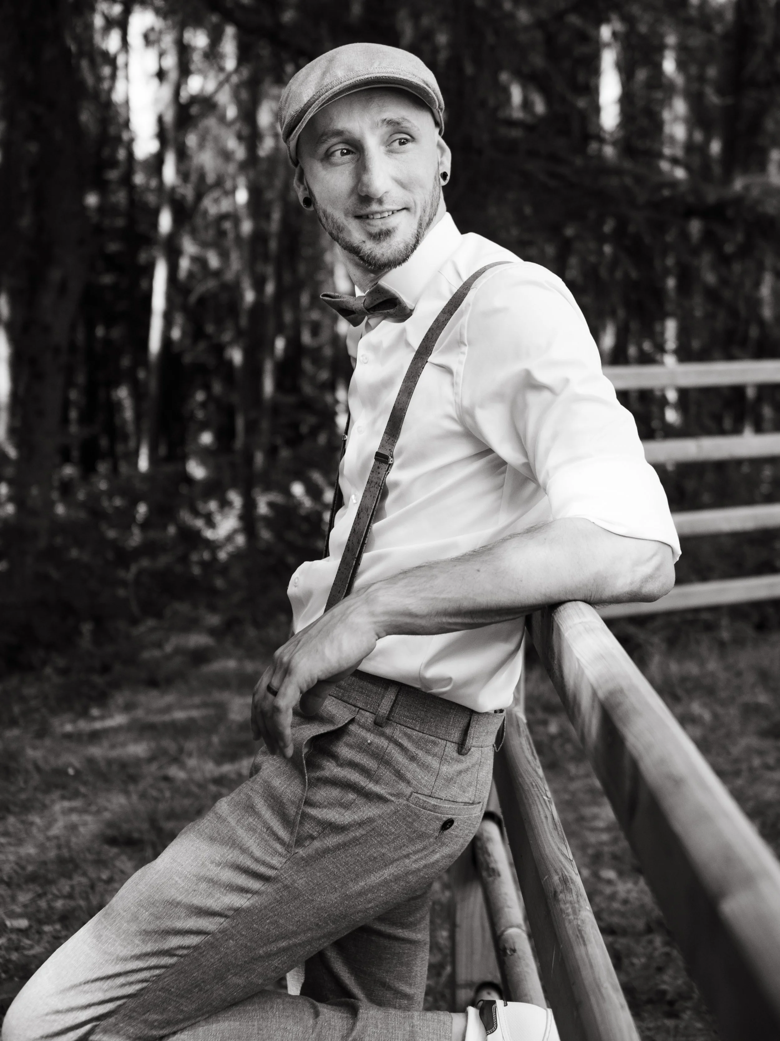 A man leaning on a wooden railing outdoors, wearing a white shirt, gray trousers, a bow tie, suspenders, a flat cap, and earrings, smiling and looking to the side.