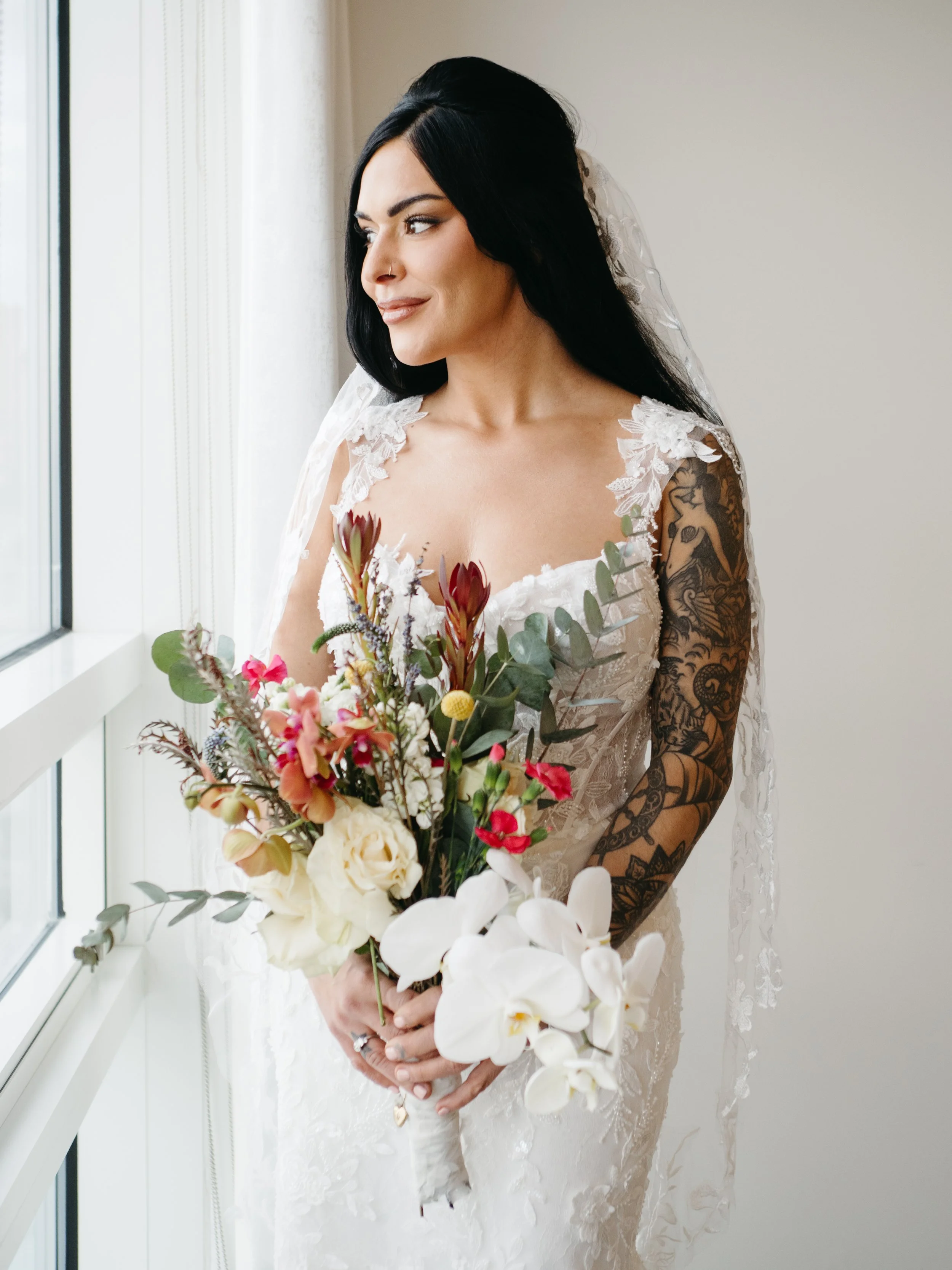 A woman in a white wedding dress holding a bouquet, standing by a window with natural light, with black hair and tattoos on her right arm.