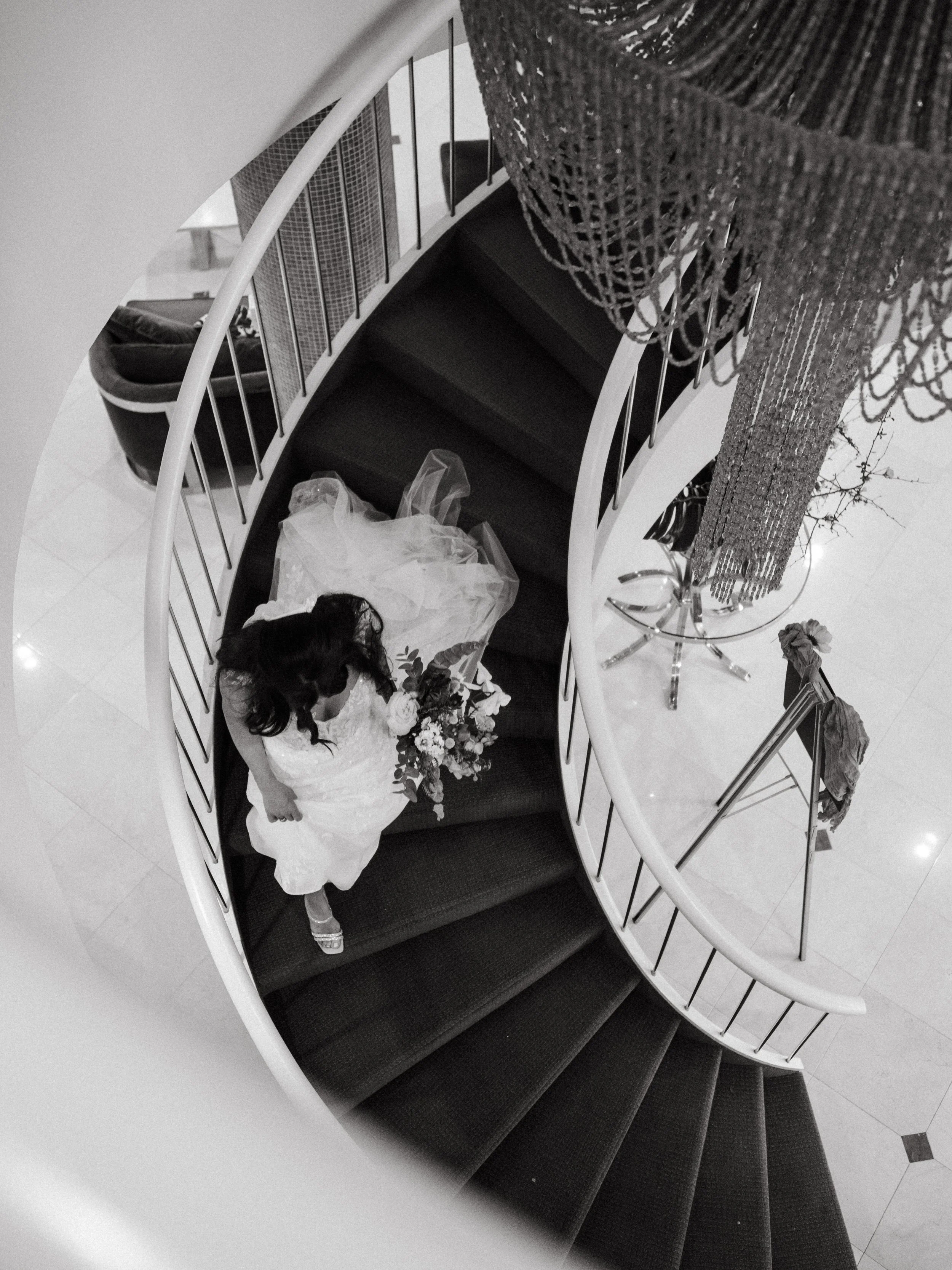 A woman in a wedding dress holding a bouquet standing on a curved staircase, viewed from above.