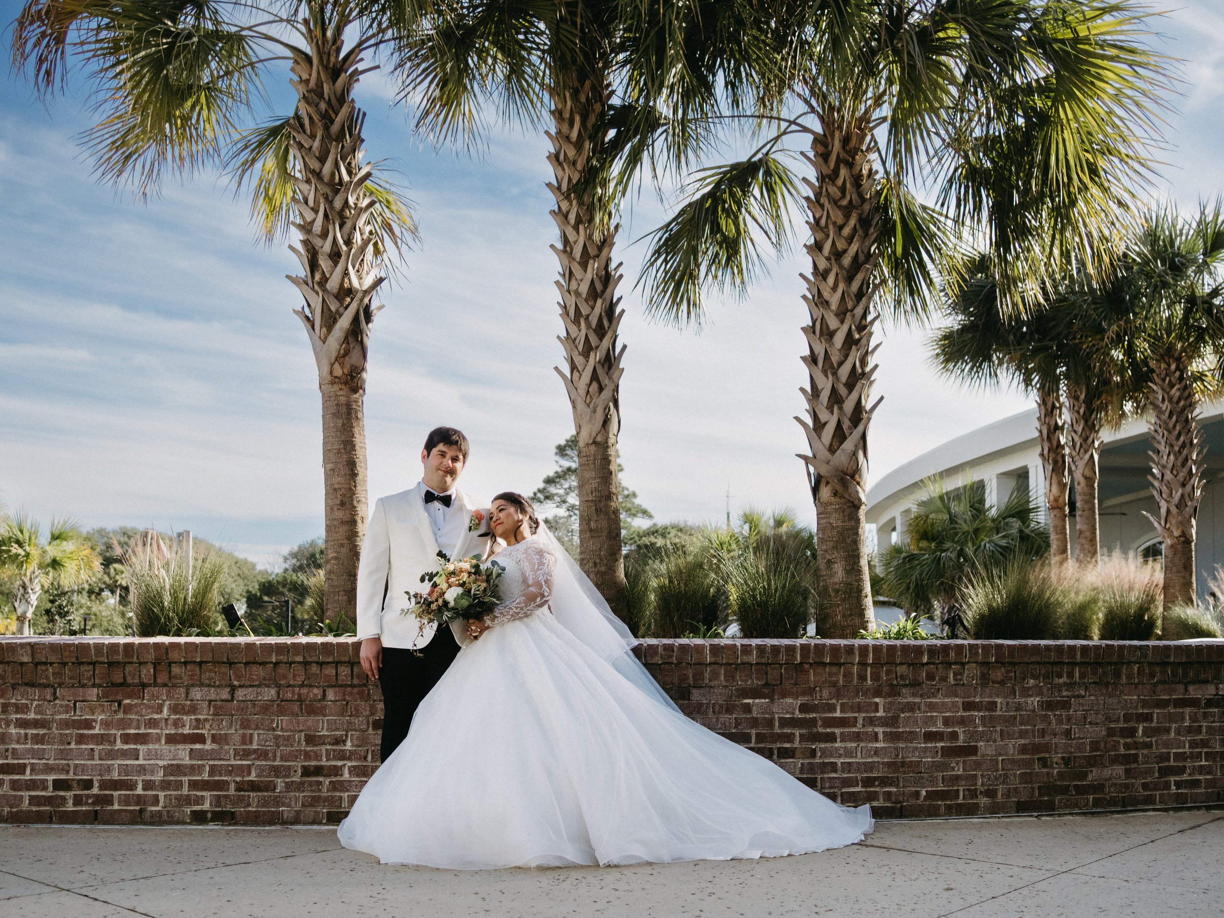 A bride and groom dressed in wedding attire standing outdoors in front of palm trees and a brick wall.