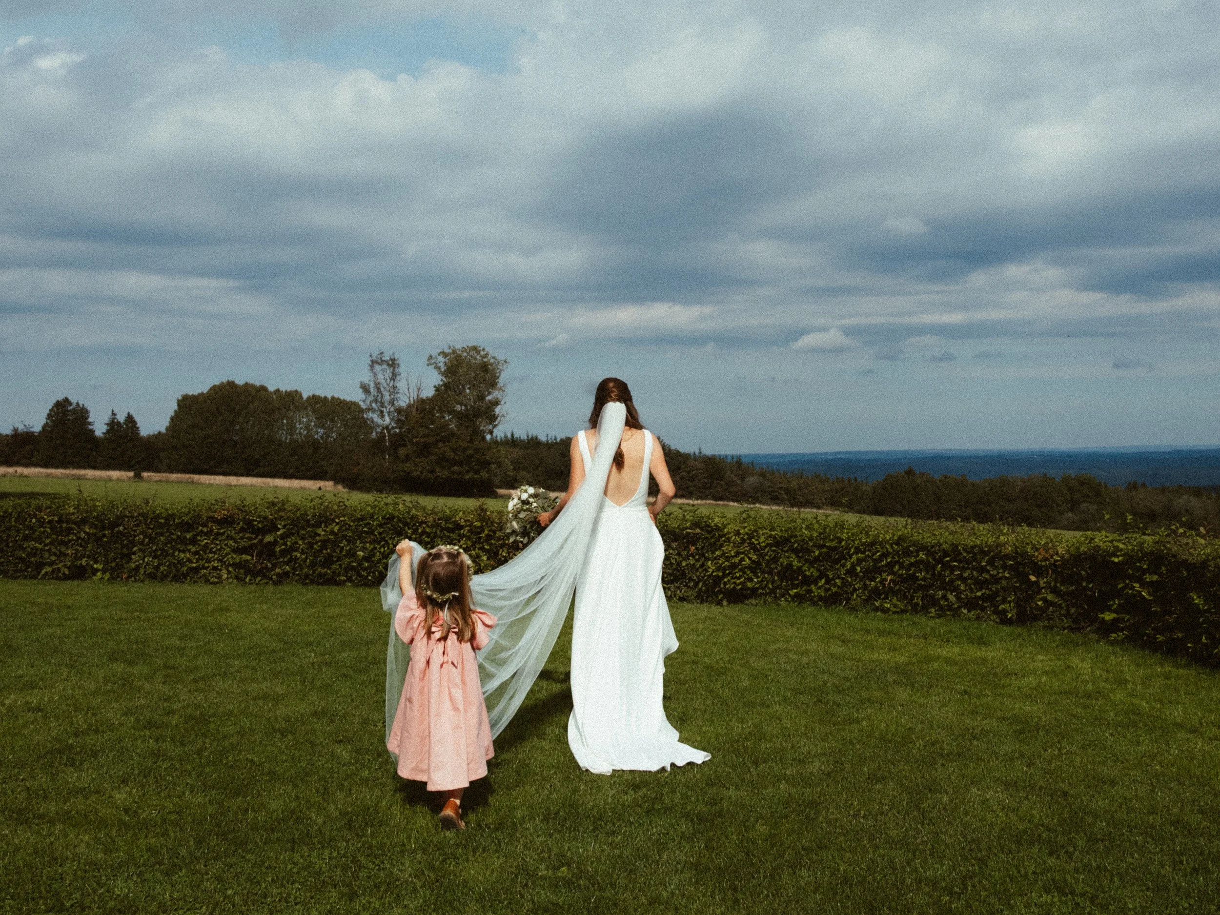 A woman in a white wedding dress holding a bouquet of flowers walking on a grassy field with a young girl in a pink dress, with a scenic background of trees, bushes, and a cloudy sky.