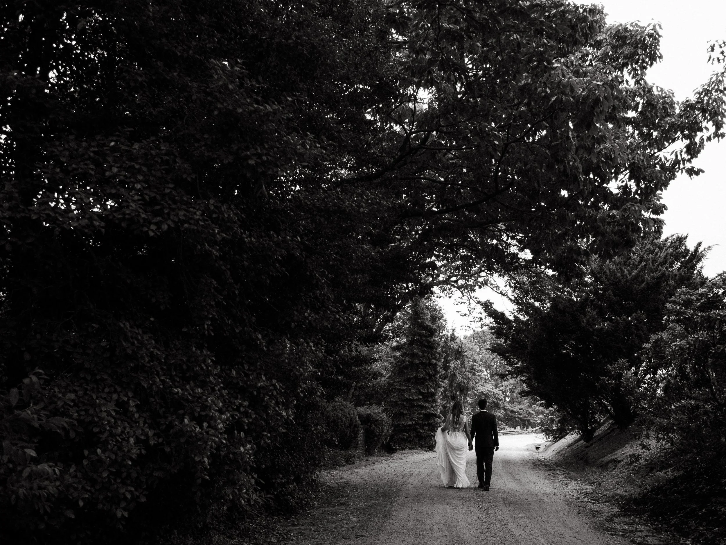 A bride and groom walking together on a dirt path surrounded by trees, viewed from behind.
