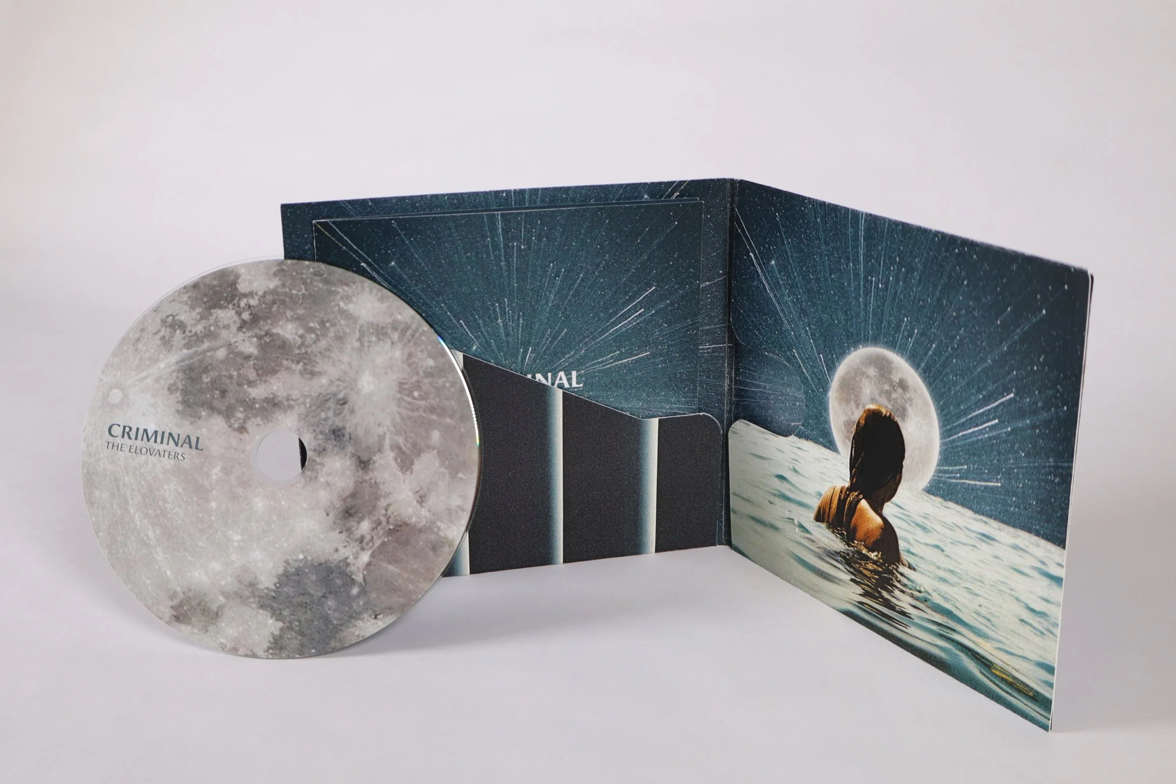 A CD titled 'Criminal' by The Elovaters, with a moon and starry sky scene, and an album booklet with a woman swimming towards a large moon in the background.