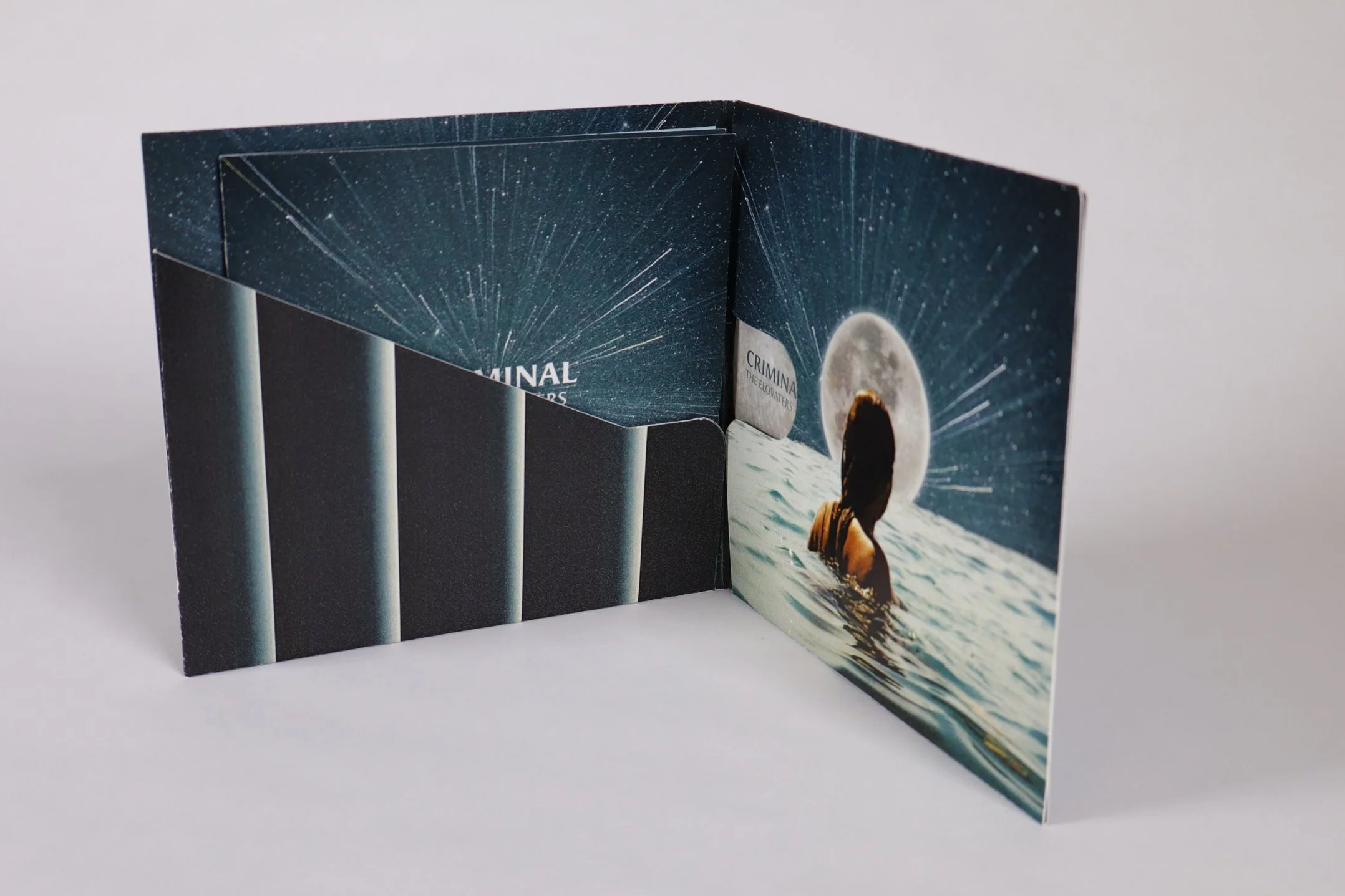 An open small booklet or card with space-themed images, featuring a woman in water with a large moon and starry sky in the background, labeled 'CRIMINA'.