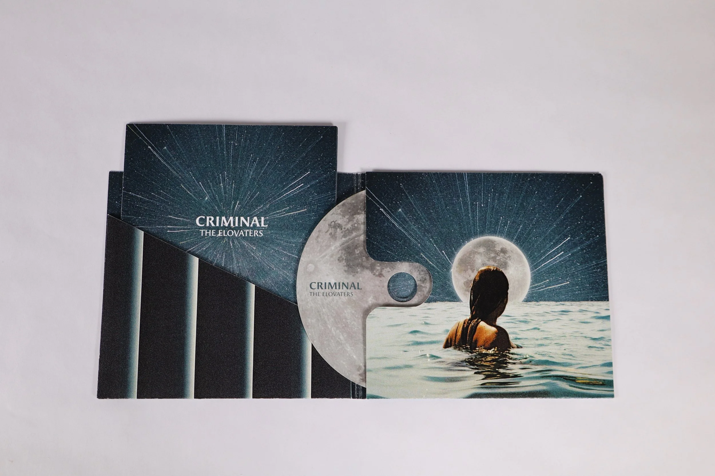 A CD case with a booklet inside, labeled 'CRIMINAL THE ELOVATERS,' featuring a starry night sky and a full moon, with a person swimming in water in front of the moon.