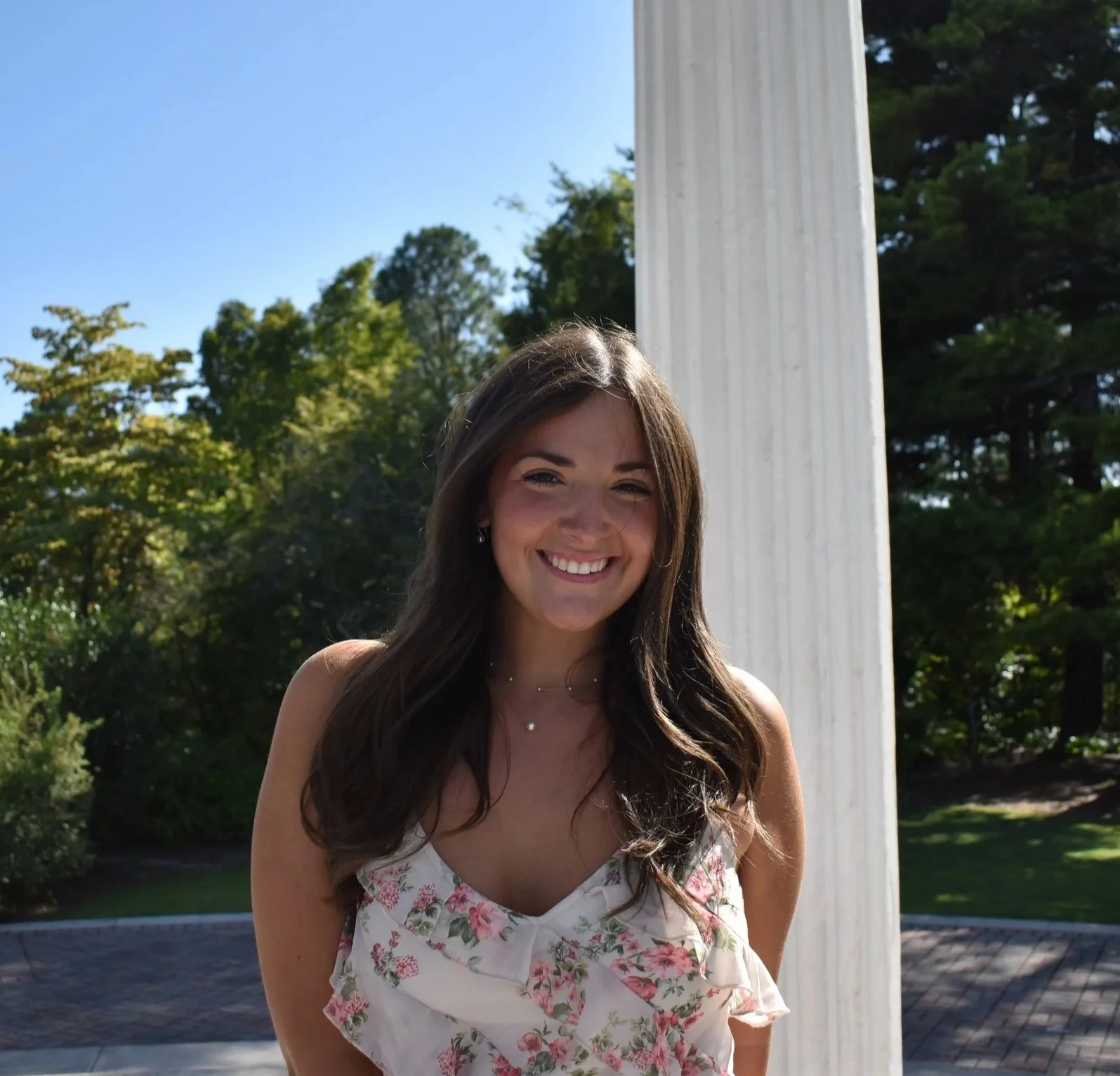 Young woman smiling outdoors next to a white column, wearing a floral top and jewelry, with green trees and a blue sky in the background.