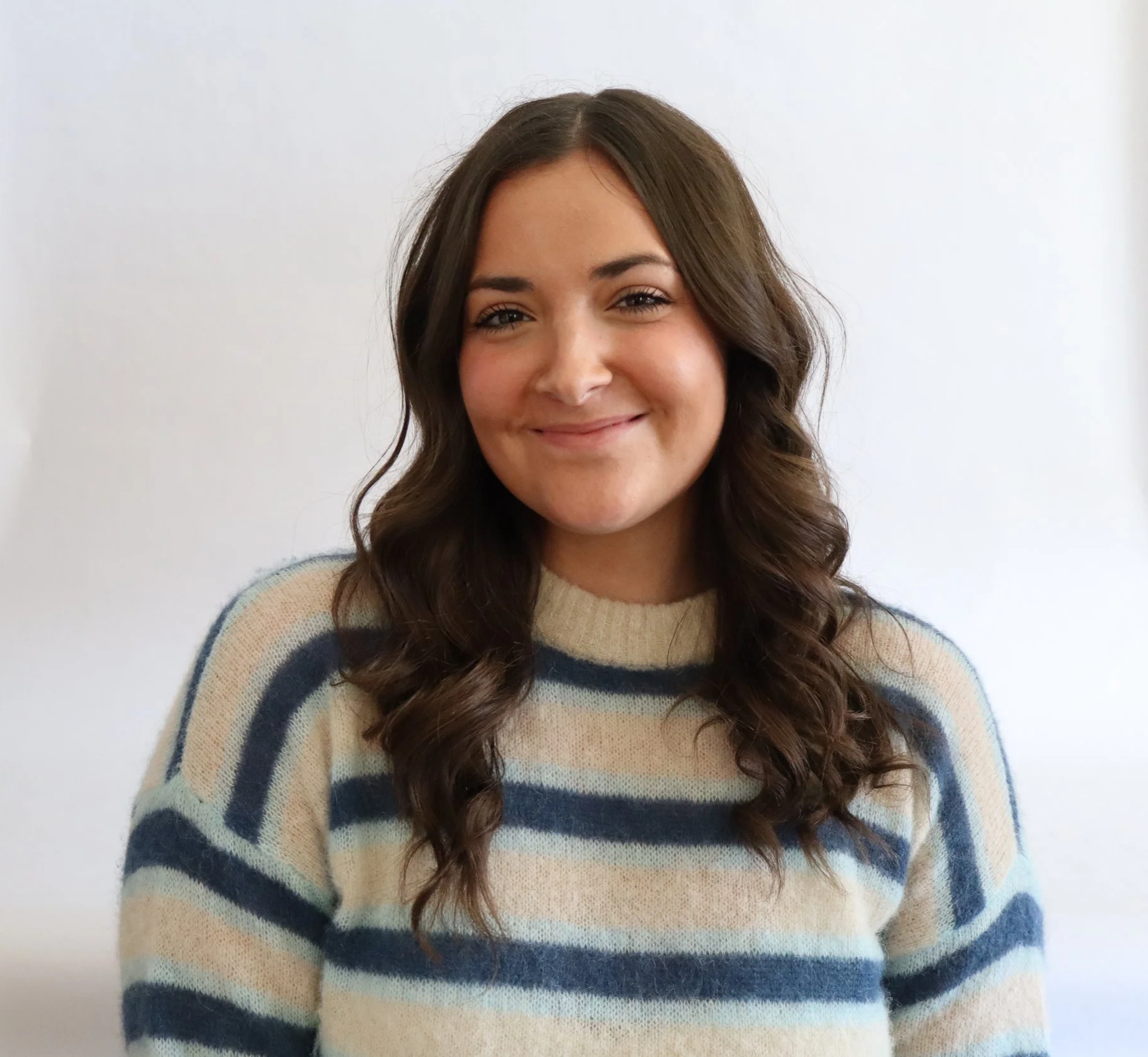 A young woman with long, wavy brown hair wearing a beige and blue striped sweater, smiling softly, standing against a plain white background.