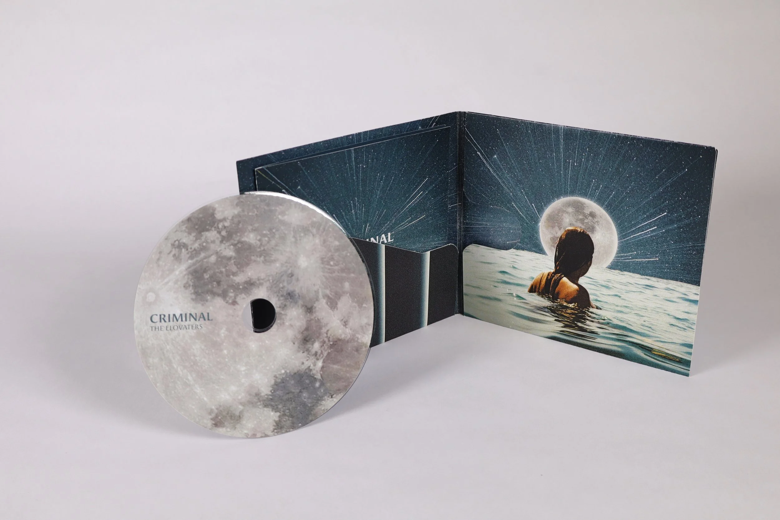 A CD titled "CRIMINAL" by THE ELOVATERS next to an open album cover with a woman in water, starry sky, and a full moon.