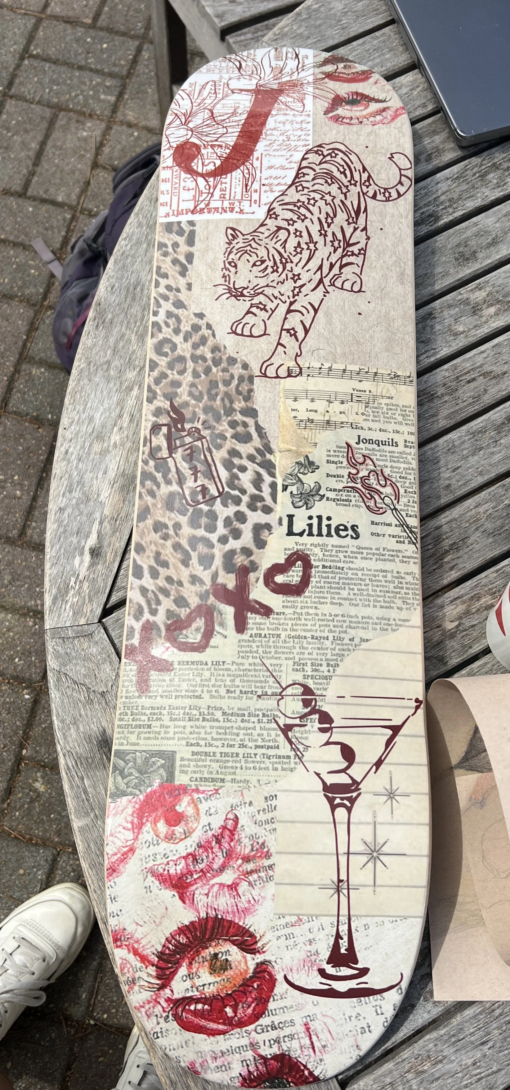 Decorated skateboard deck with various doodles and cutout images, including a tiger, eye, leopard print, flowers, lips, and handwritten text, placed on a wooden bench next to a smartphone and a drink.