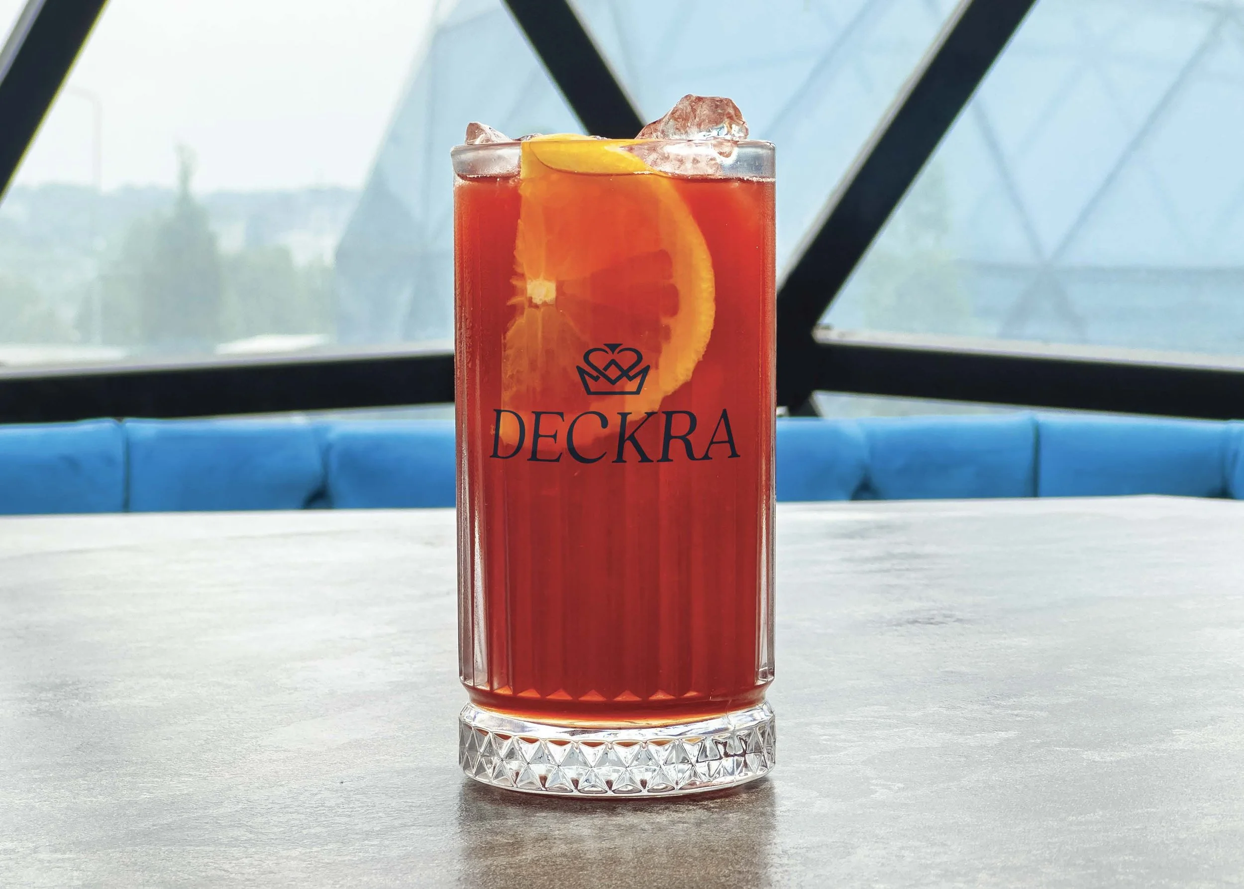 A tall glass of red iced beverage with orange slices and ice cubes, labeled 'Deckra.'