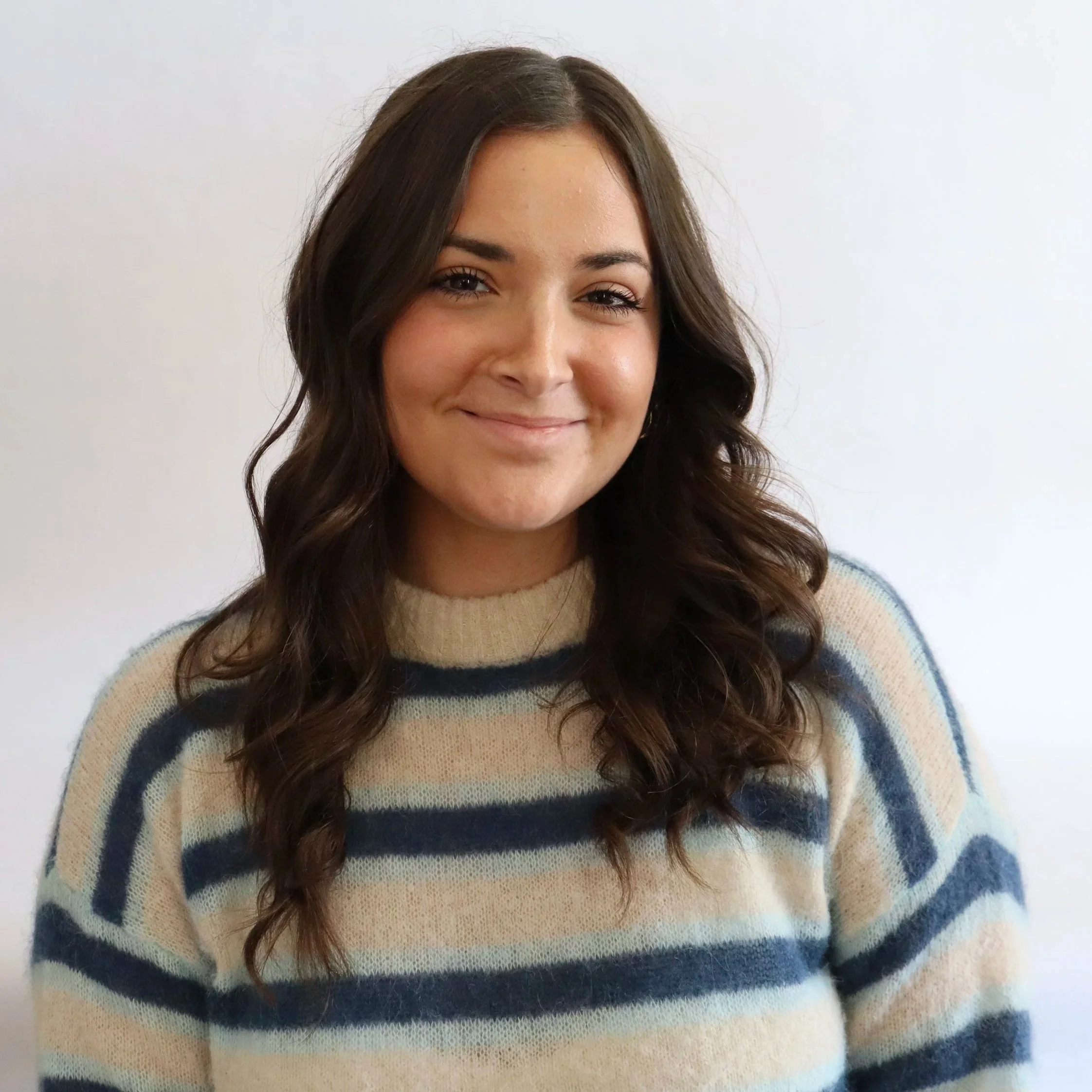 Portrait of a young woman with long brown wavy hair wearing a beige and dark blue striped sweater, smiling softly against a plain white background.