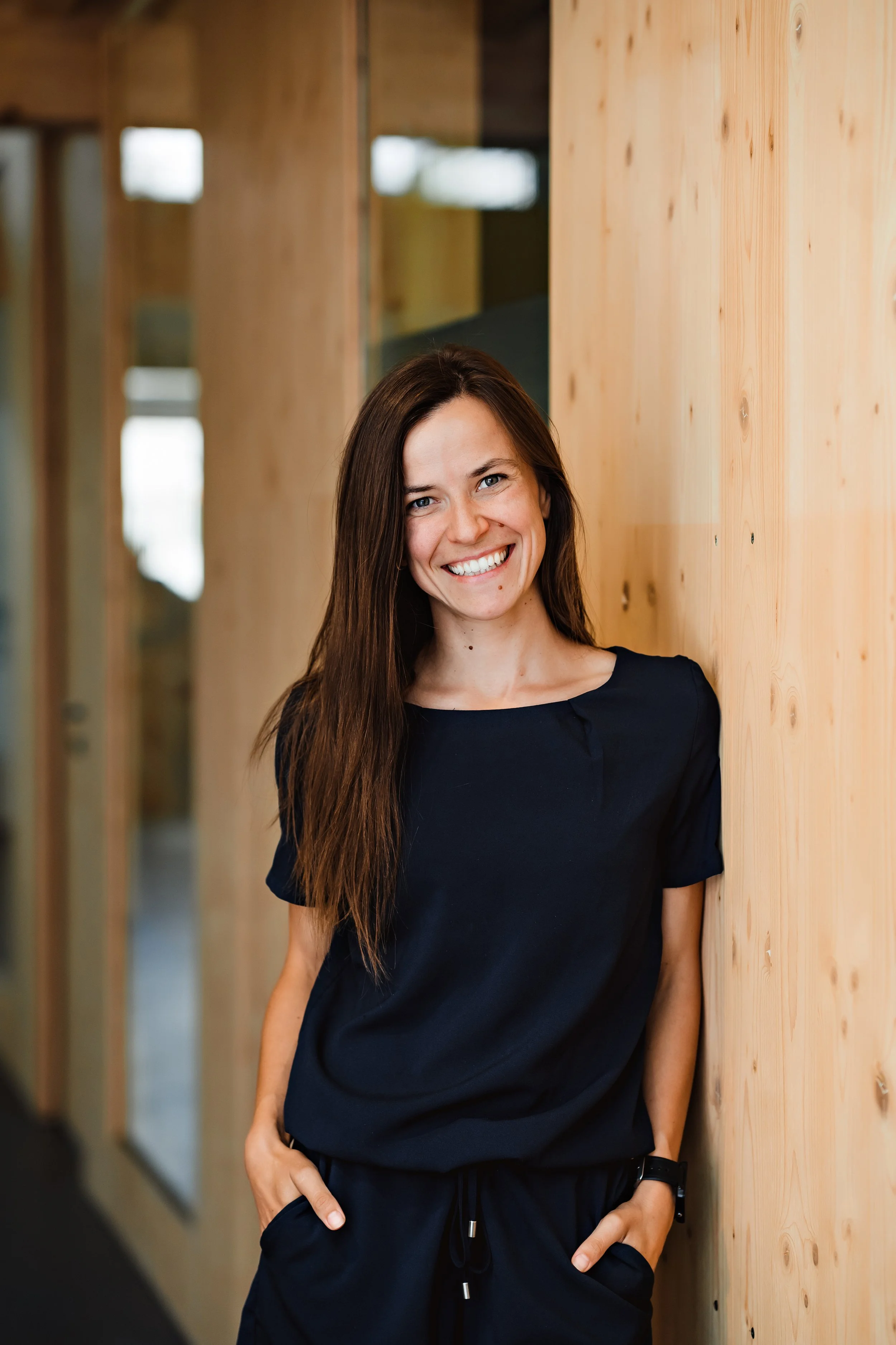 A woman with long brown hair smiling, wearing a black casual outfit, standing in front of a wooden wall and glass door with reflections.
