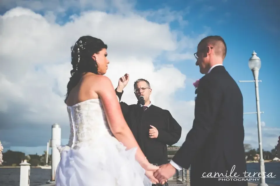 Bride and groom holding hands during wedding ceremony outdoors, officiant speaking, cloudy sky in background.
