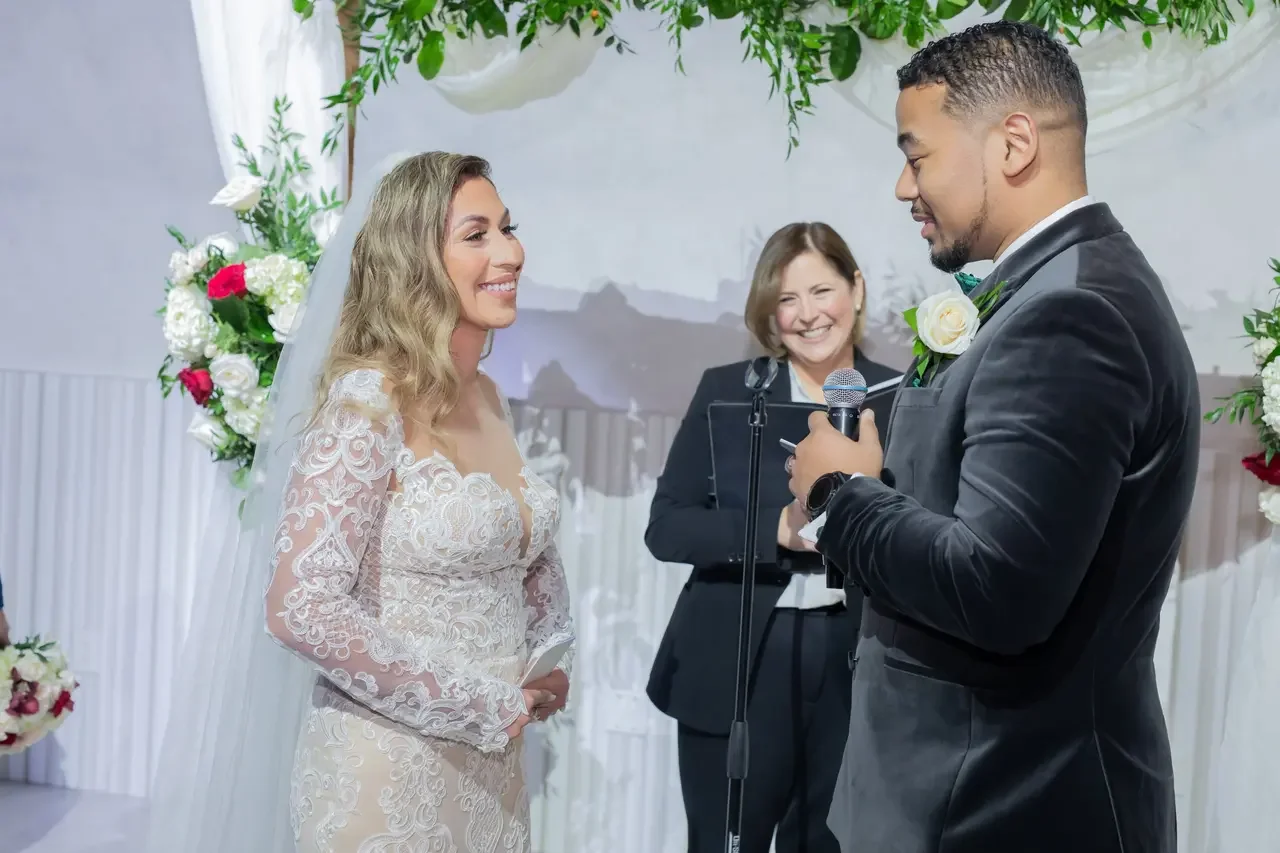 A bride and groom exchanging vows during their wedding ceremony, with the officiant smiling in the background. The bride is wearing a lace wedding dress and the groom is in a black tuxedo, both smiling at each other. The setting is decorated with flowers and greenery.