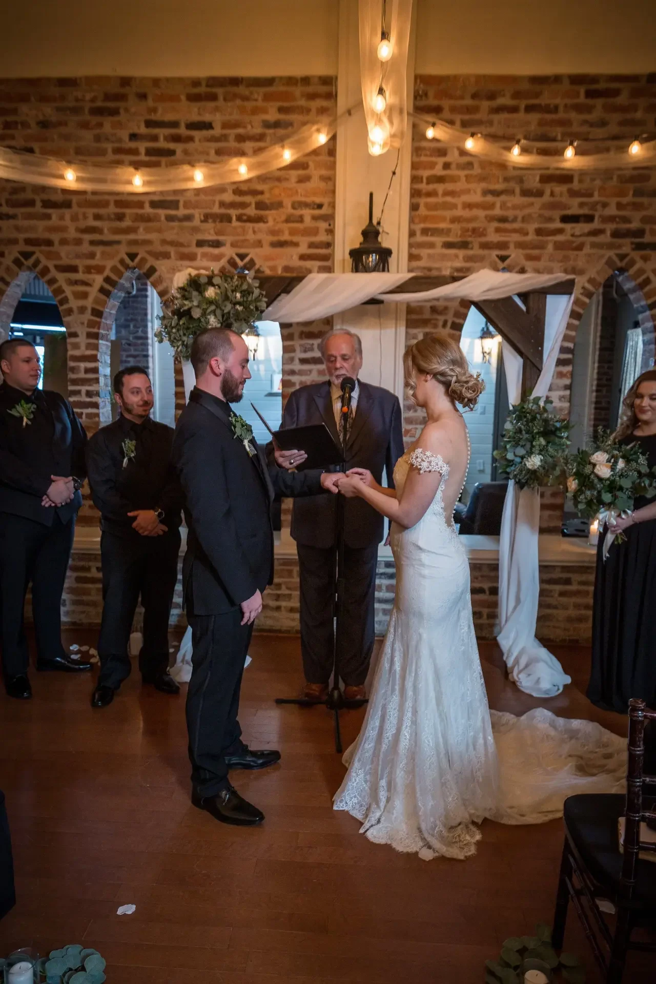 A bride and groom exchanging vows during their wedding ceremony in a rustic indoor venue with brick walls and string lights overhead. The officiant stands behind them, with bridesmaids and groomsmen on either side, all dressed formally.