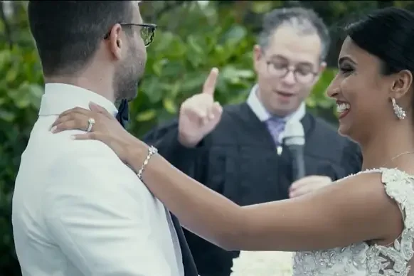 A bride and groom exchanging vows during a wedding ceremony outdoors, with an officiant speaking in the background.