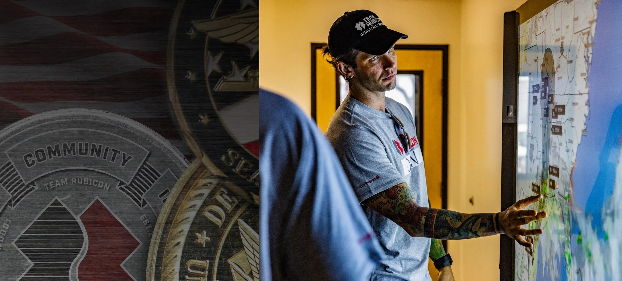 A man with tattoos on his right arm, wearing a gray T-shirt with the 'Team Rubicon' logo and a black cap, is pointing at a large digital map or display with a stylized map and markers. Another person, partially visible with a denim shirt, is nearby. The setting appears to be an indoor conference or office.