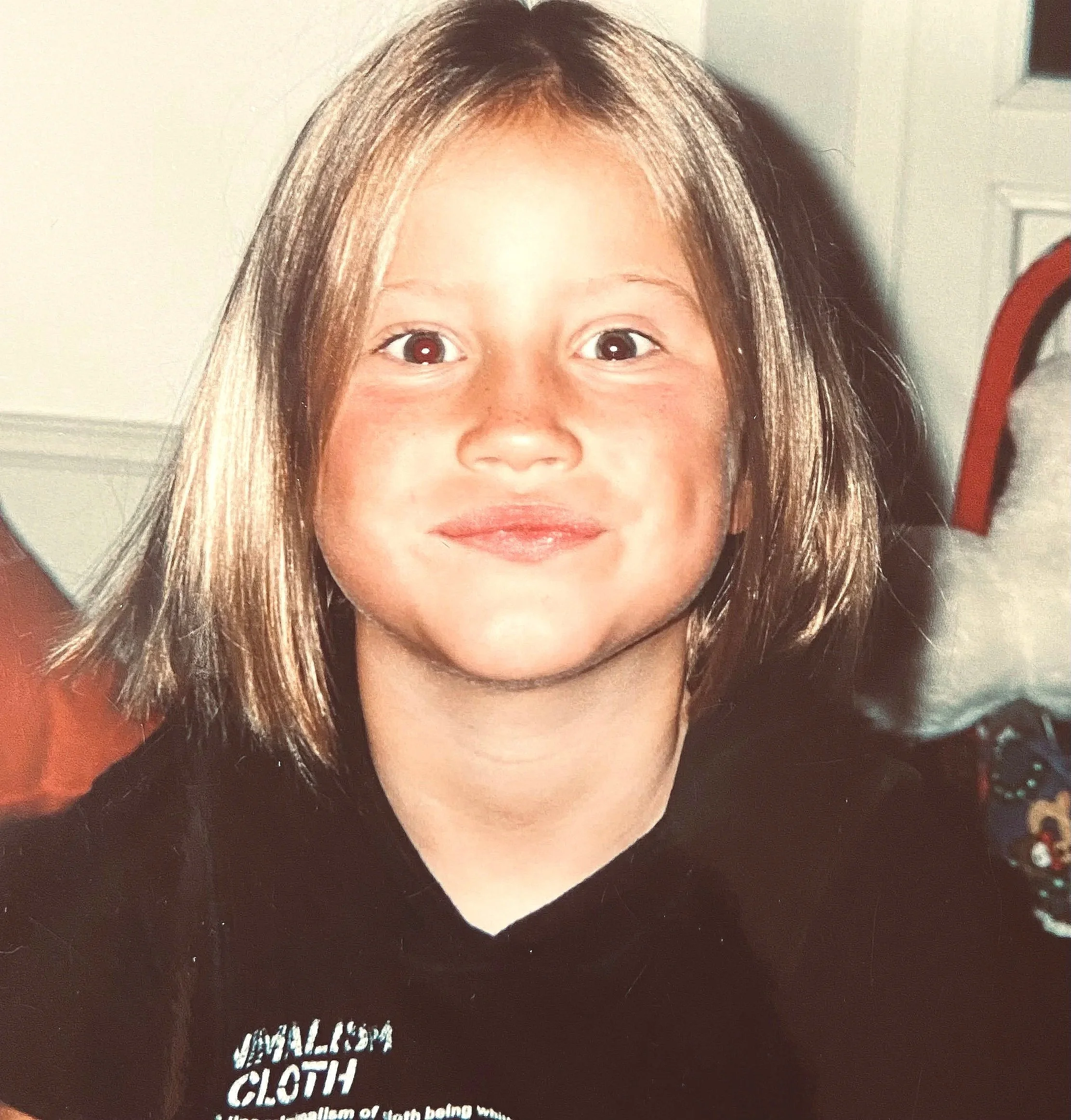 Jeune fille aux cheveux bruns courts, souriant, portant un t-shirt noir et assise dans une pièce intérieure.