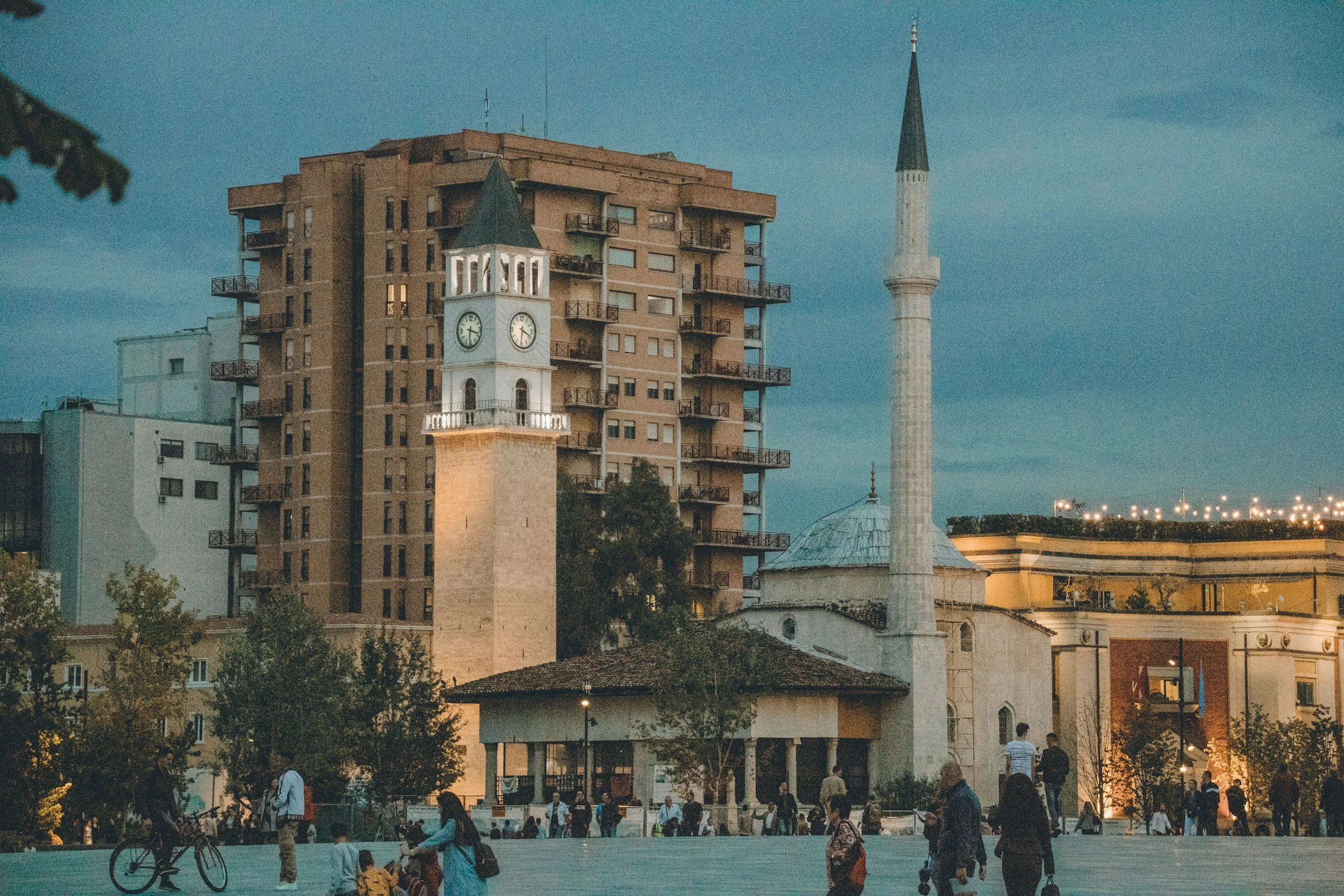 Cityscape at dusk featuring a tall modern building, a clock tower, and a mosque minaret with people walking and cycling in the foreground.