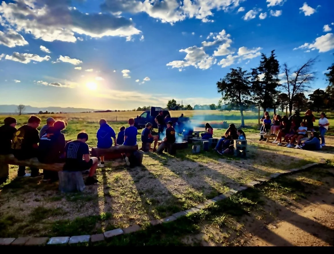 A group of people gathered outdoors in a rural setting during sunset, sitting on benches and steps, near a barbecue grill, with trees and open fields in the background.