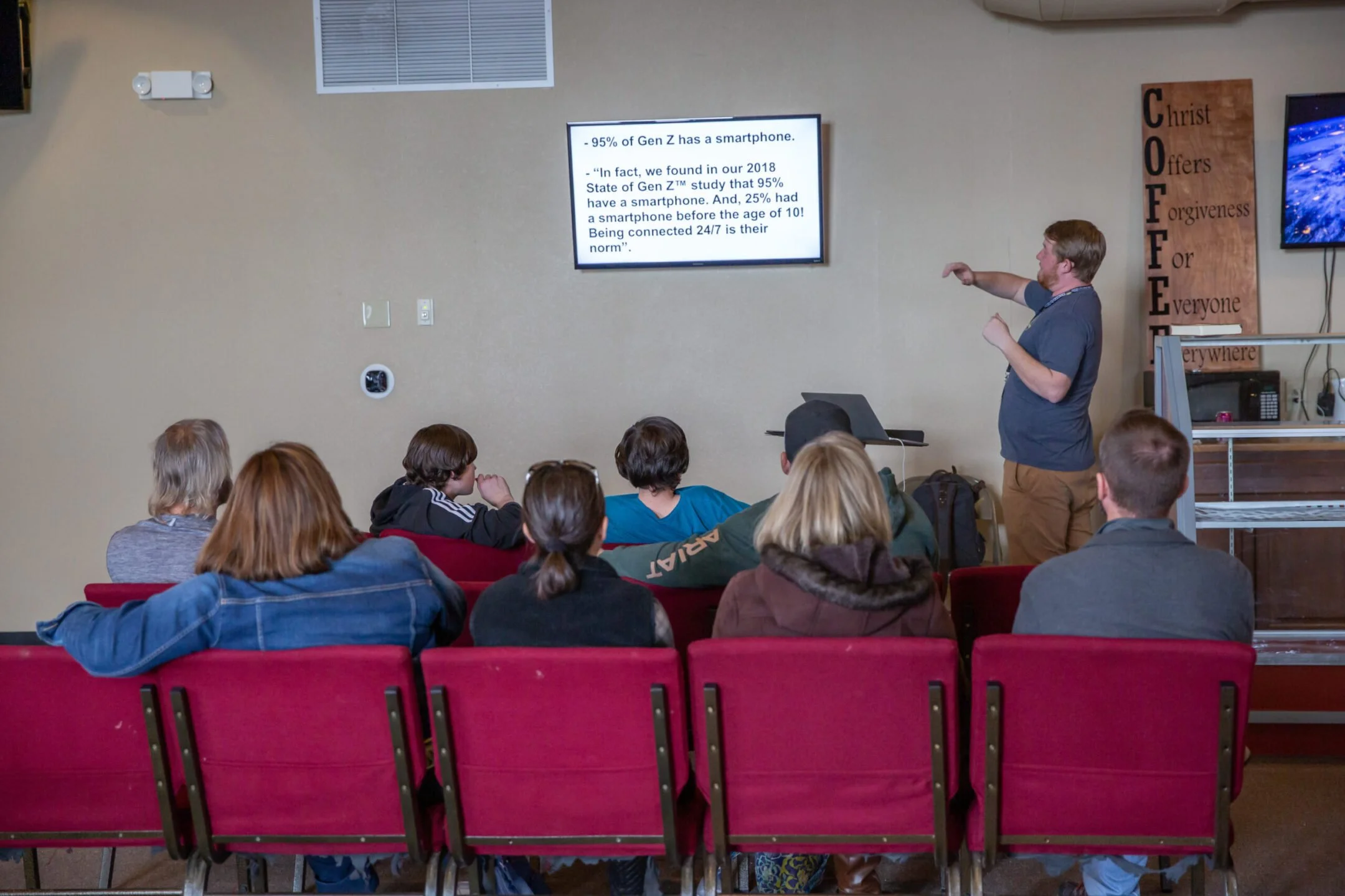 A man giving a presentation to an audience in a room with red chairs. The presenter stands near a large screen displaying a slide with statistical information about smartphones. The audience includes young people, some wearing hoodies and jackets, facing the presenter and screen.