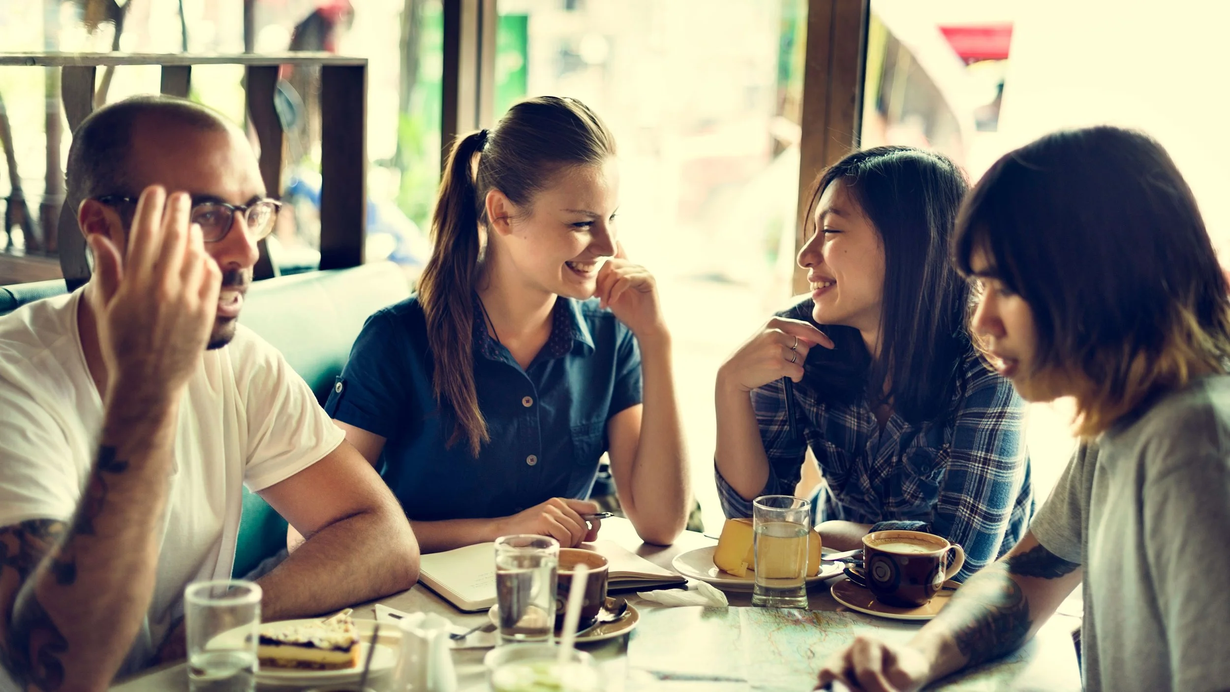 Group of four people sitting at a table in a cafe, engaging in conversation and laughing, with drinks and desserts on the table.