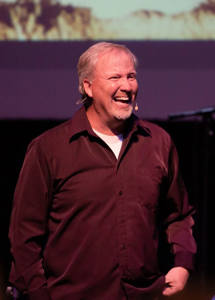 Man smiling and laughing on stage with a dark background and indistinct scenery.