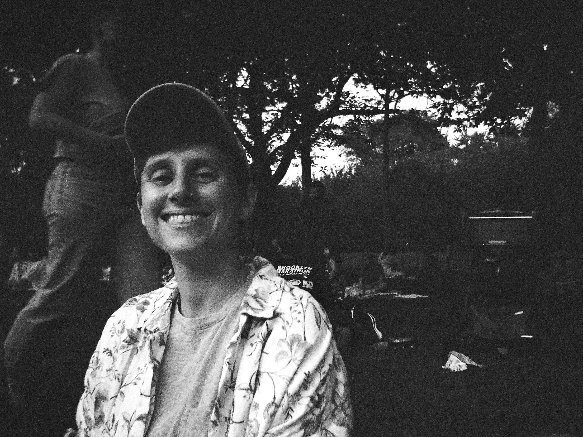 A grainy, black & white photo of a smiling person wearing a baseball cap and floral shirt outdoors at dusk, with trees and a people walking in the background.