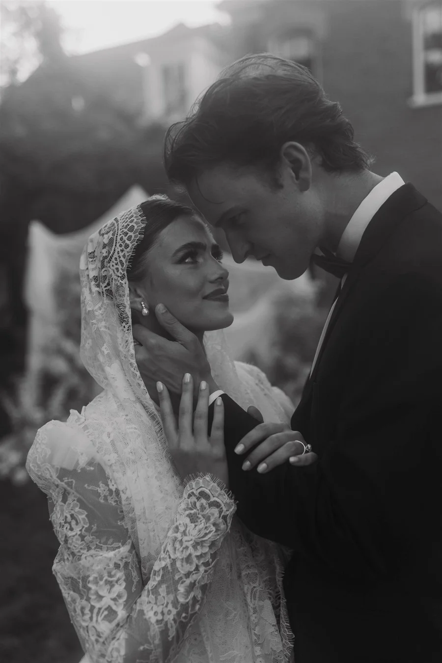 Black and white photo of a bride and groom in an intimate moment, with their foreheads touching. The bride wears lace attire and a lace veil, while the groom is dressed in a tuxedo with a bow tie, outdoors with blurred background of trees and buildings.