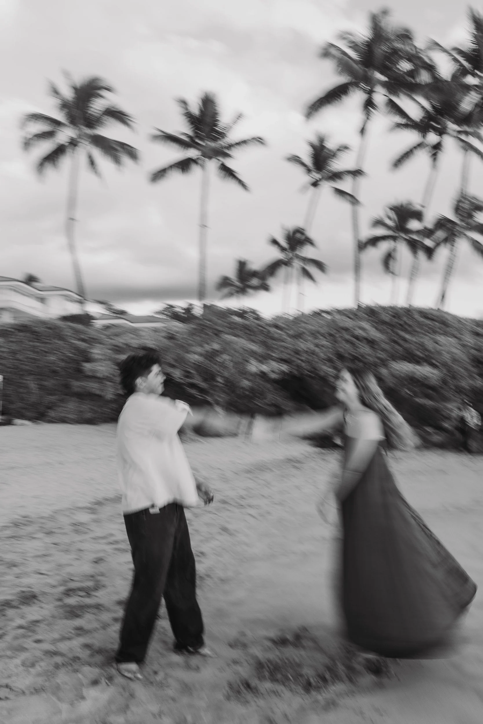 A black and white photo of a man and a woman on a beach, reaching out to each other with smiling expressions, with palm trees and cloudy sky in the background.