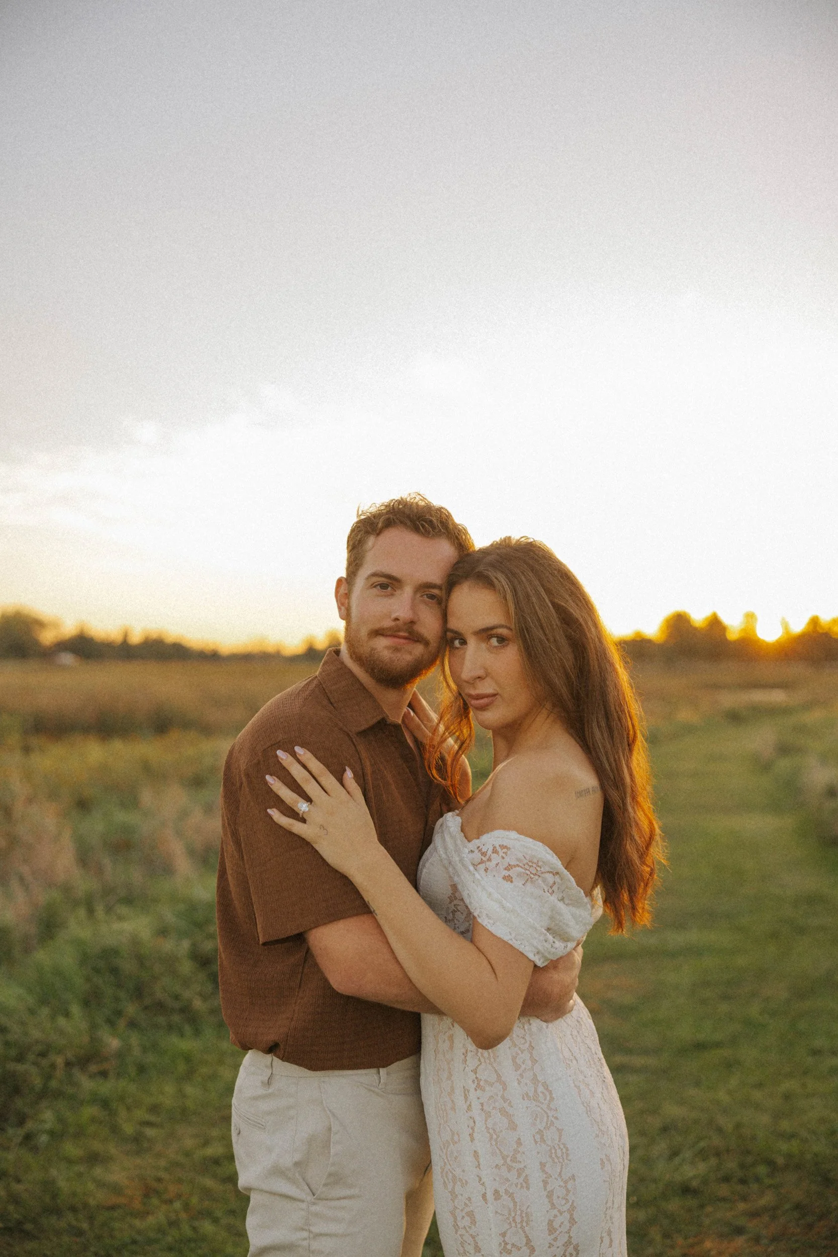 A couple embracing outdoors at sunset, with the woman wearing a white lace dress and the man wearing a brown shirt, standing on a grassy field.