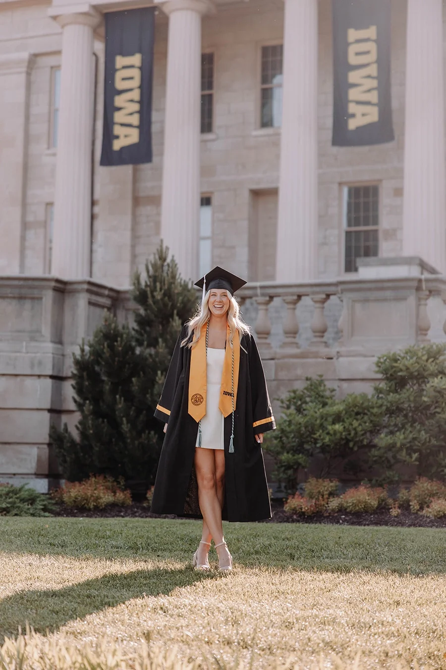 A woman in a graduation gown and cap smiling outdoors on a grassy area in front of an old stone building with large columns and banners that say 'IOWA'.