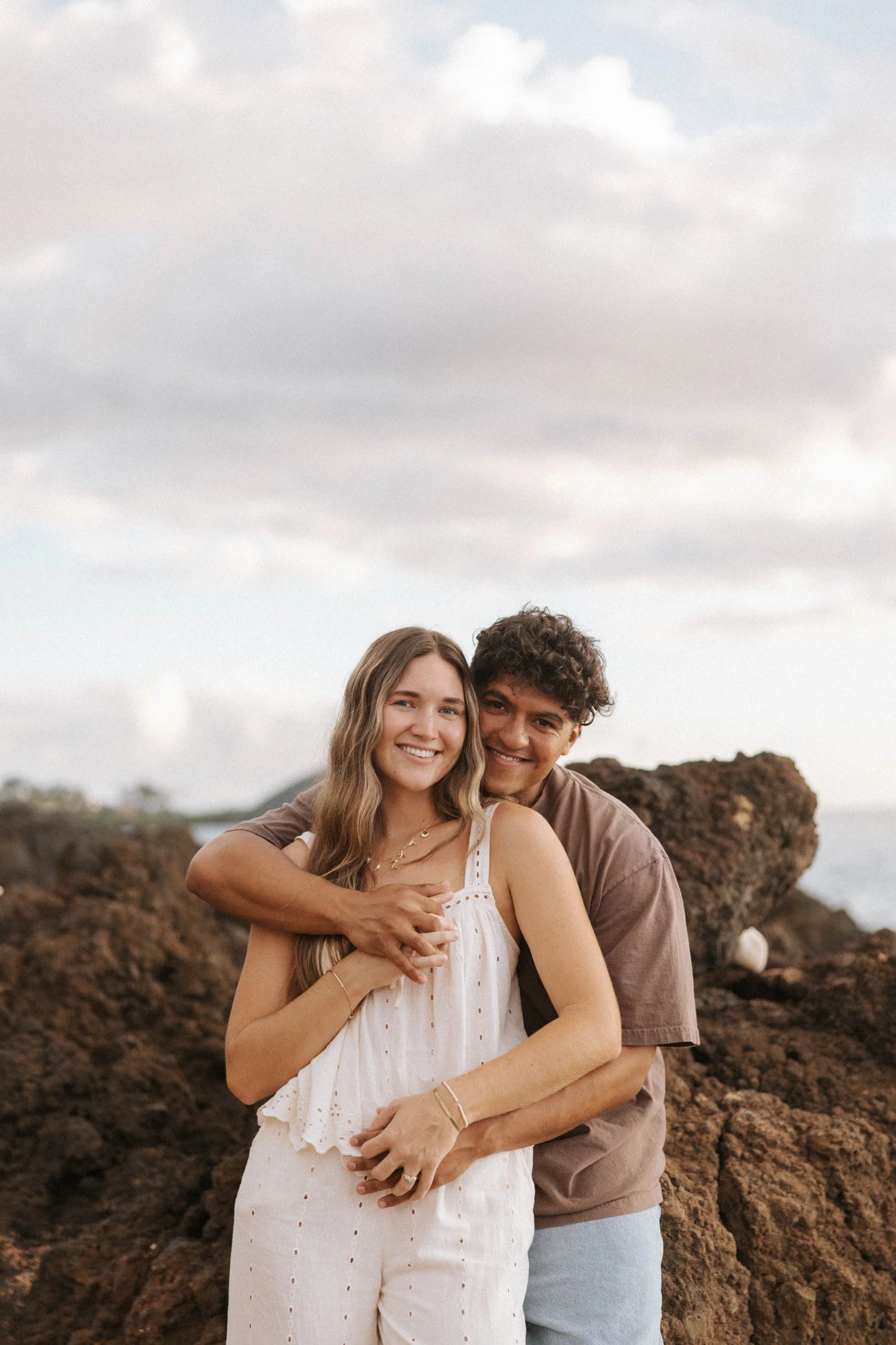 A happy couple embracing on rocky terrain outdoors, with a cloudy sky in the background.