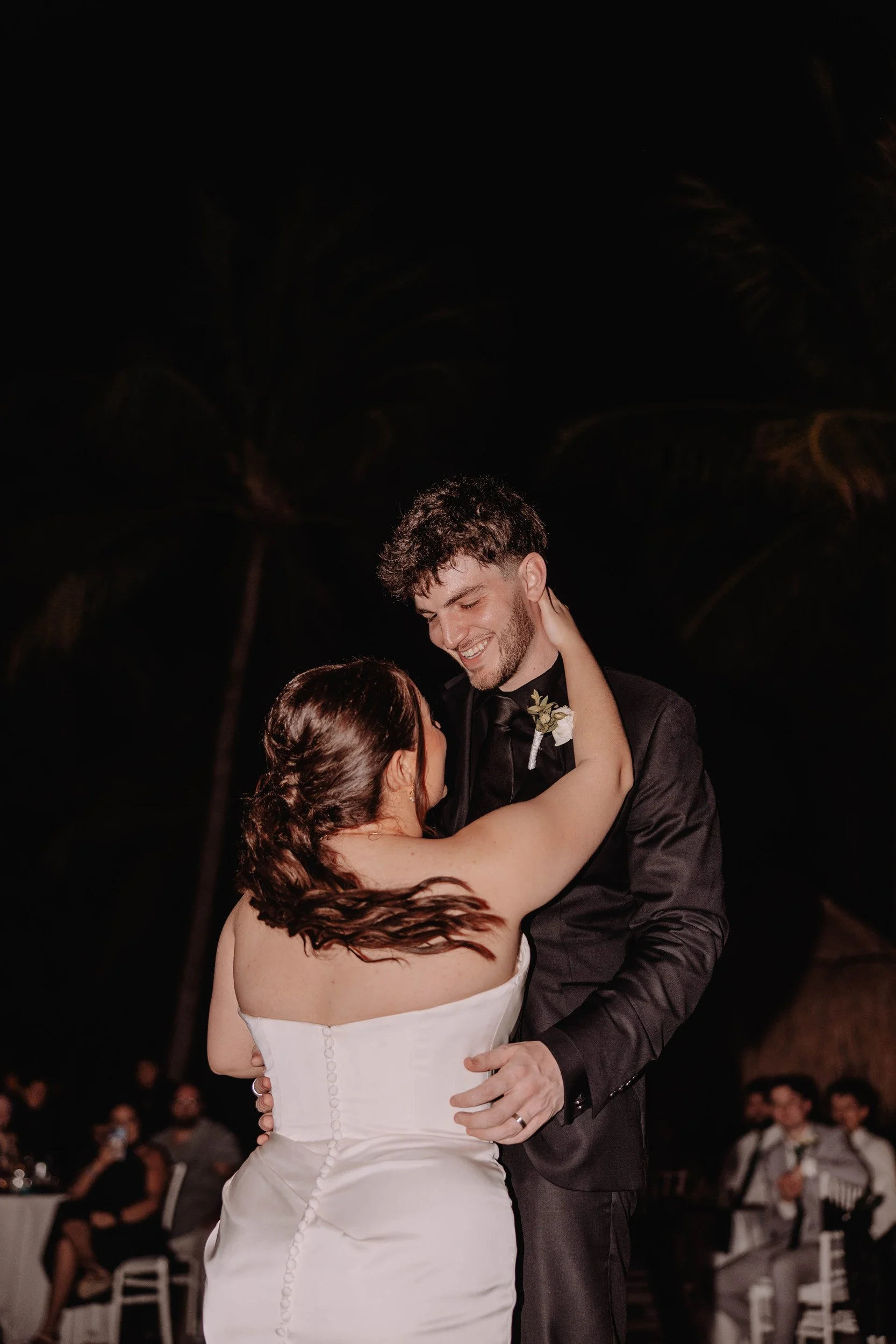 A newlywed couple sharing a dance at night, with the bride in a strapless white dress and the groom in a black suit, surrounded by guests and palm trees.