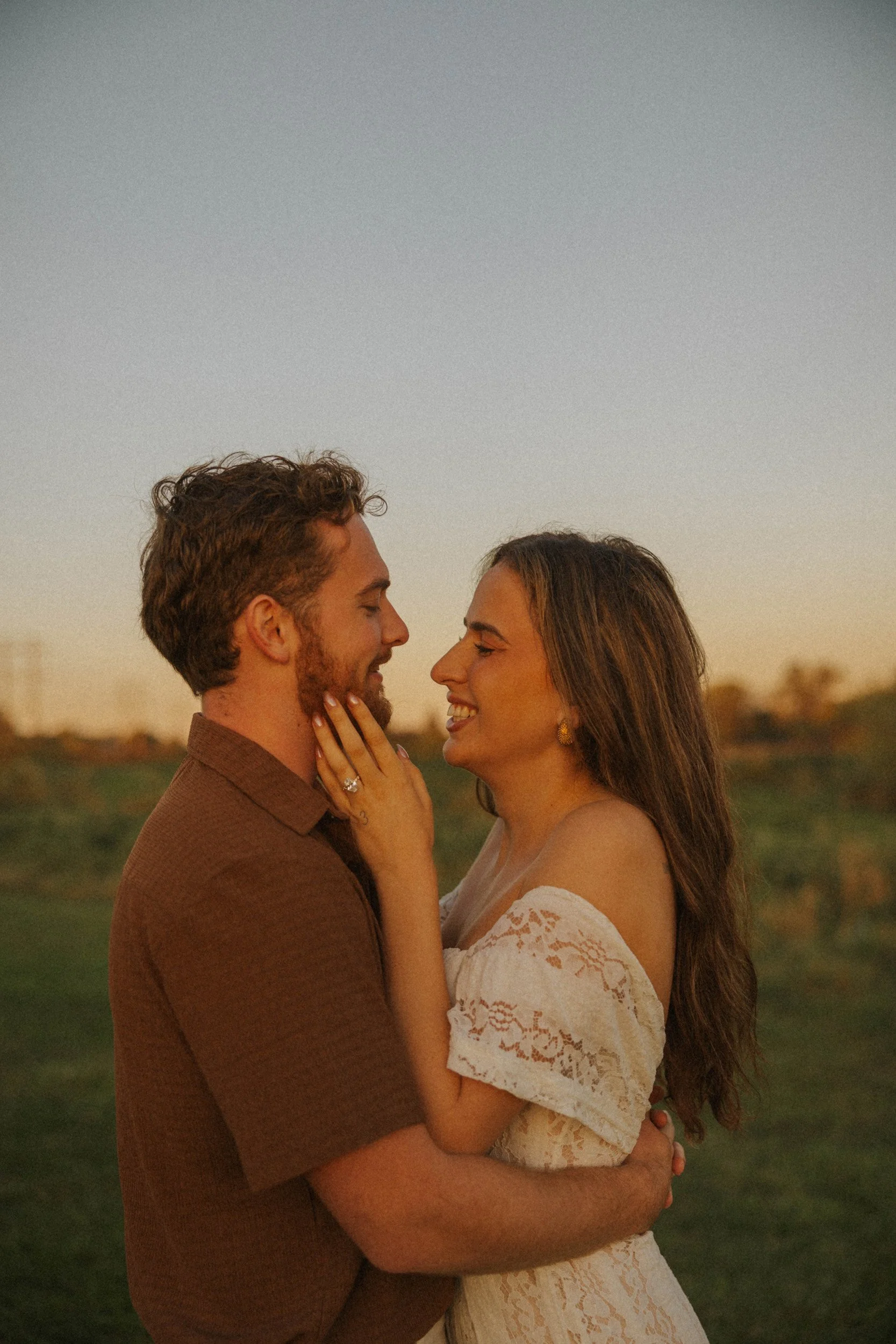 A couple in love smiles and embraces each other outdoors at sunset, with the woman touching the man's face.