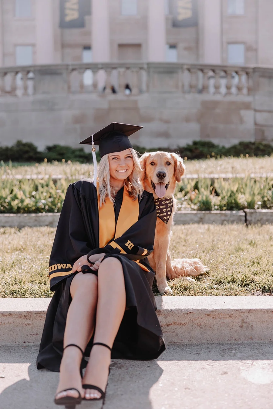A young woman in a graduation cap and gown sitting on a sidewalk with her golden retriever, both facing the camera, outside a large academic building.