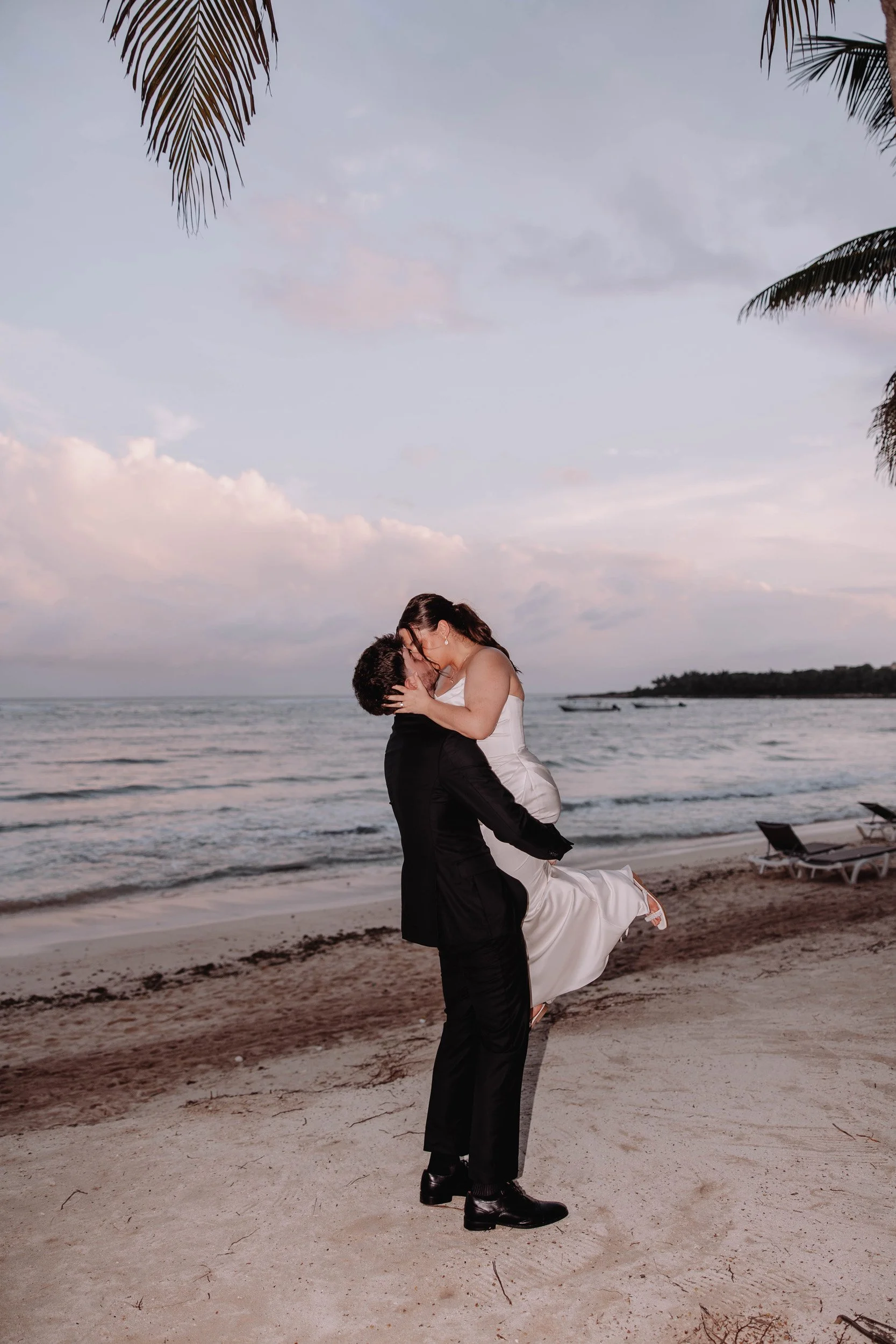 A couple dressed in wedding attire sharing a kiss on a beach at sunset, with the ocean and sky in the background and palm trees framing the scene.